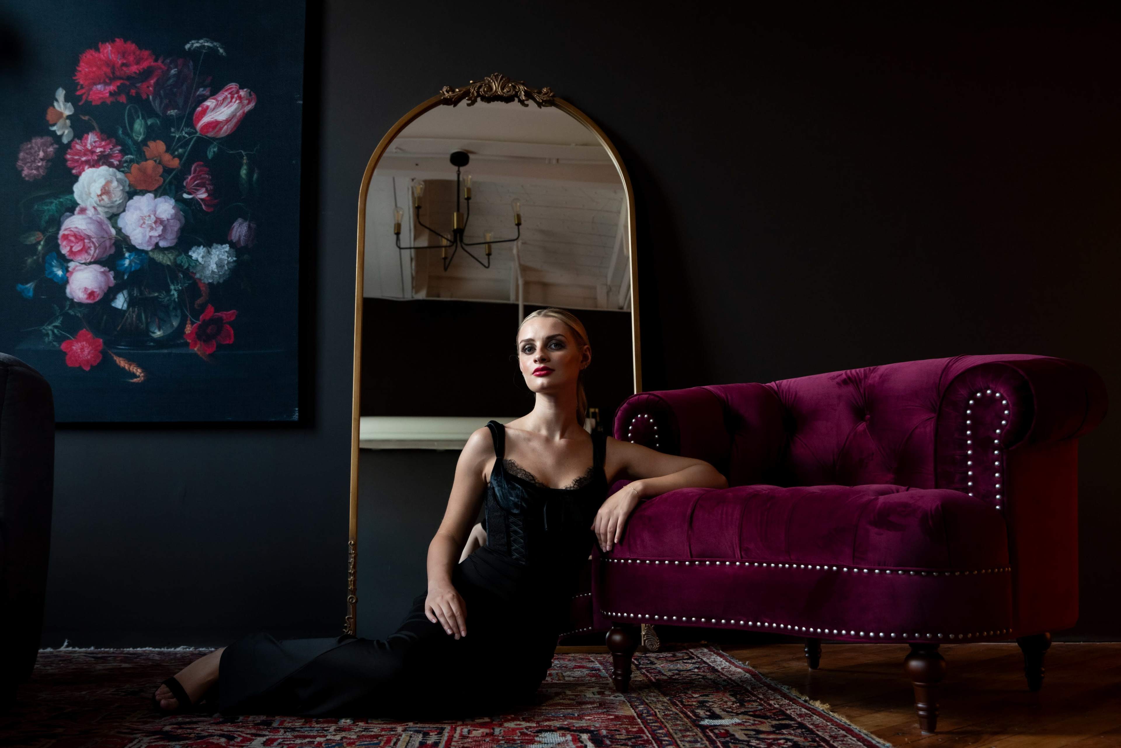 A woman in a black dress poses next to a burgundy velvet sofa, reflecting in a round mirror against a dark wall with a floral painting nearby.