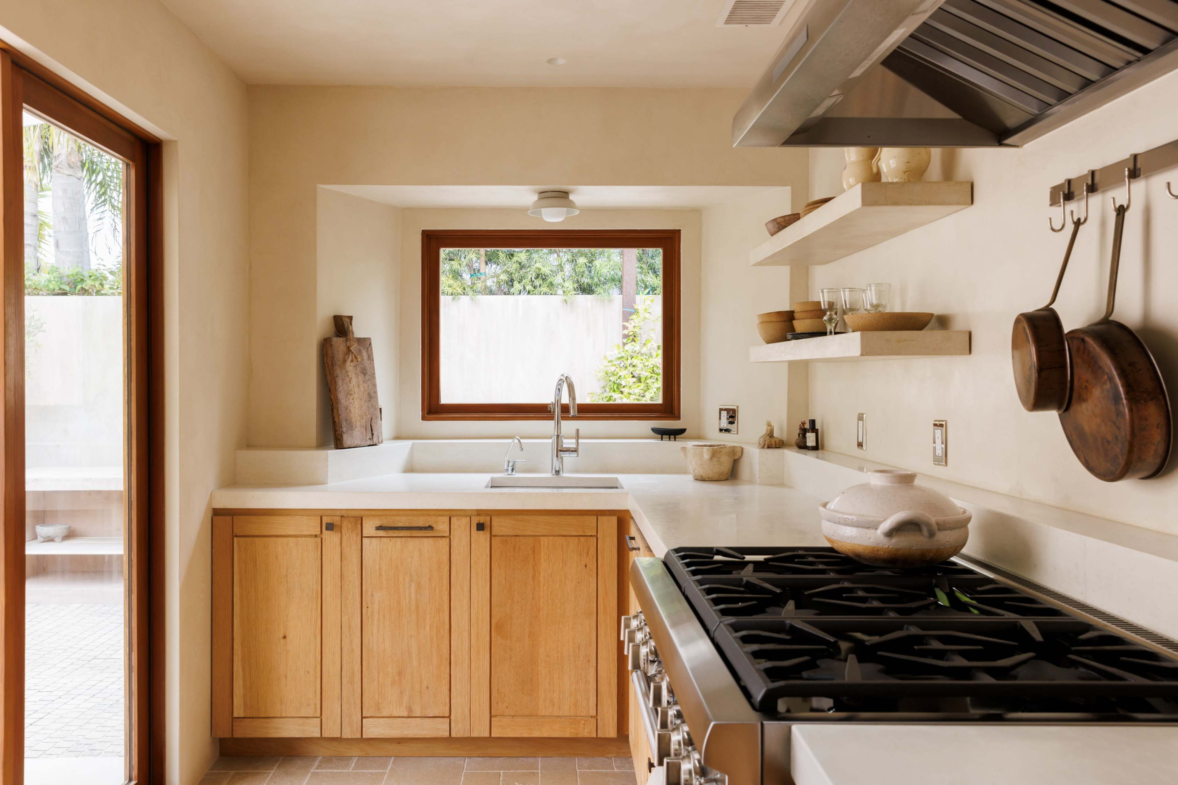 A modern kitchen features wooden cabinets, a large stovetop, and open shelves filled with cooking utensils and dishes, illuminated by natural light from a nearby window.