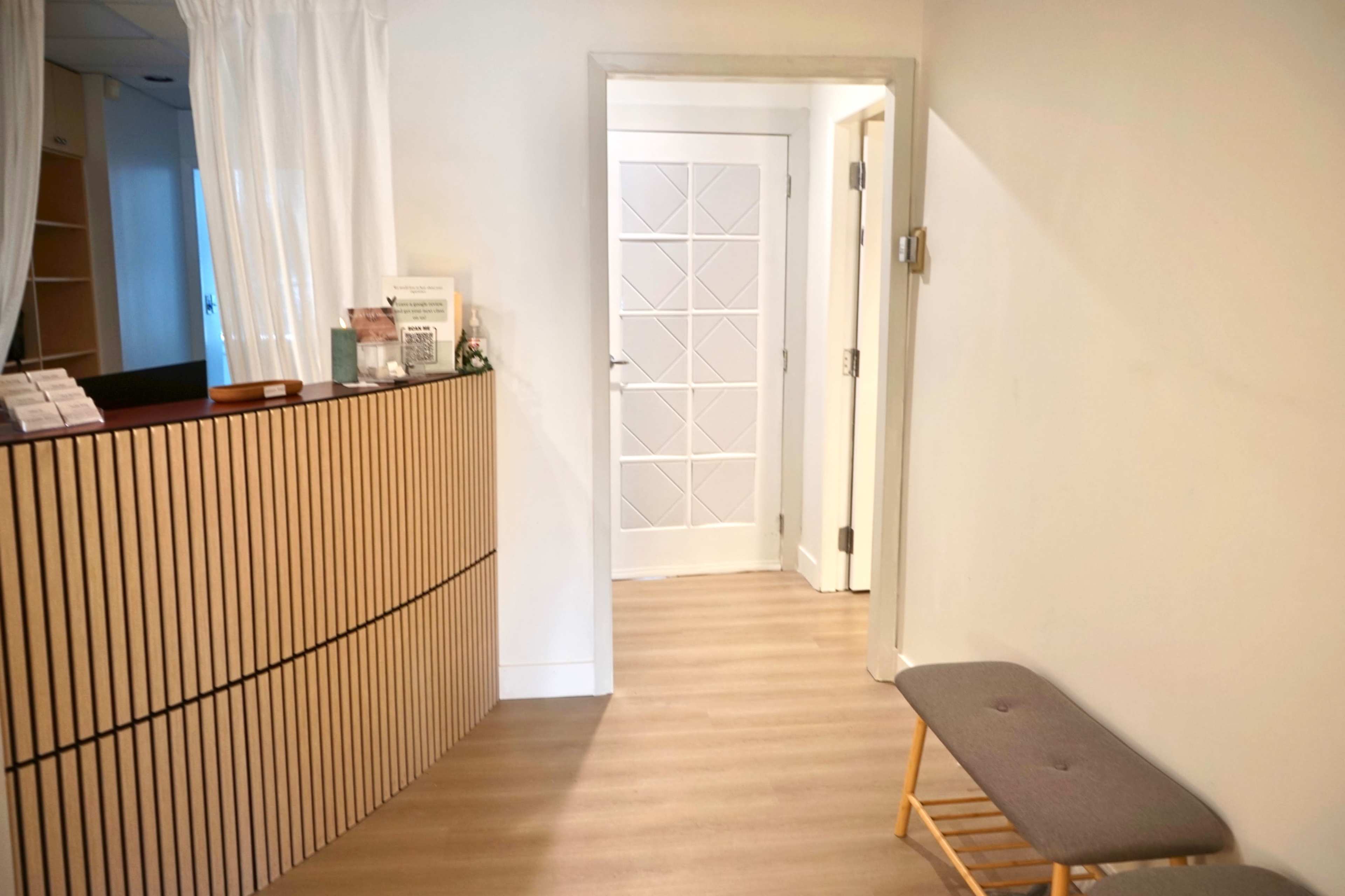 The image shows a minimalistic interior of a reception area with a wooden front desk, a door at the end of a corridor, and a bench against the wall.