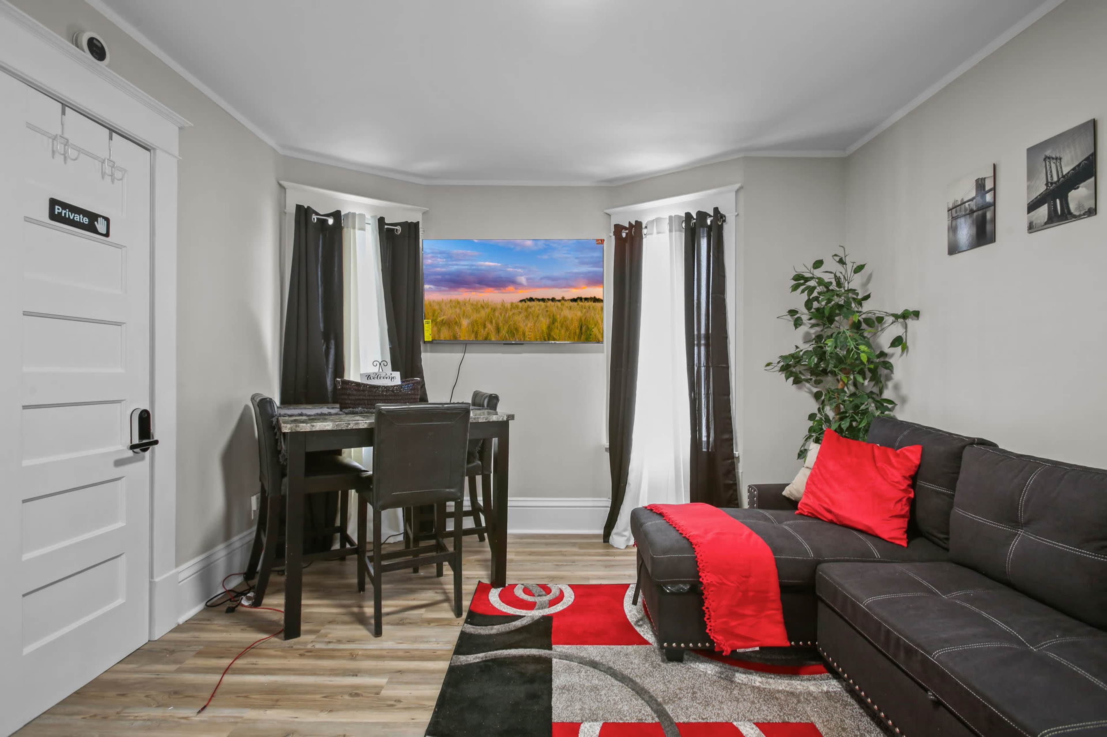 A small, modern living space features a black sectional sofa, a dining table with chairs, and a colorful area rug.