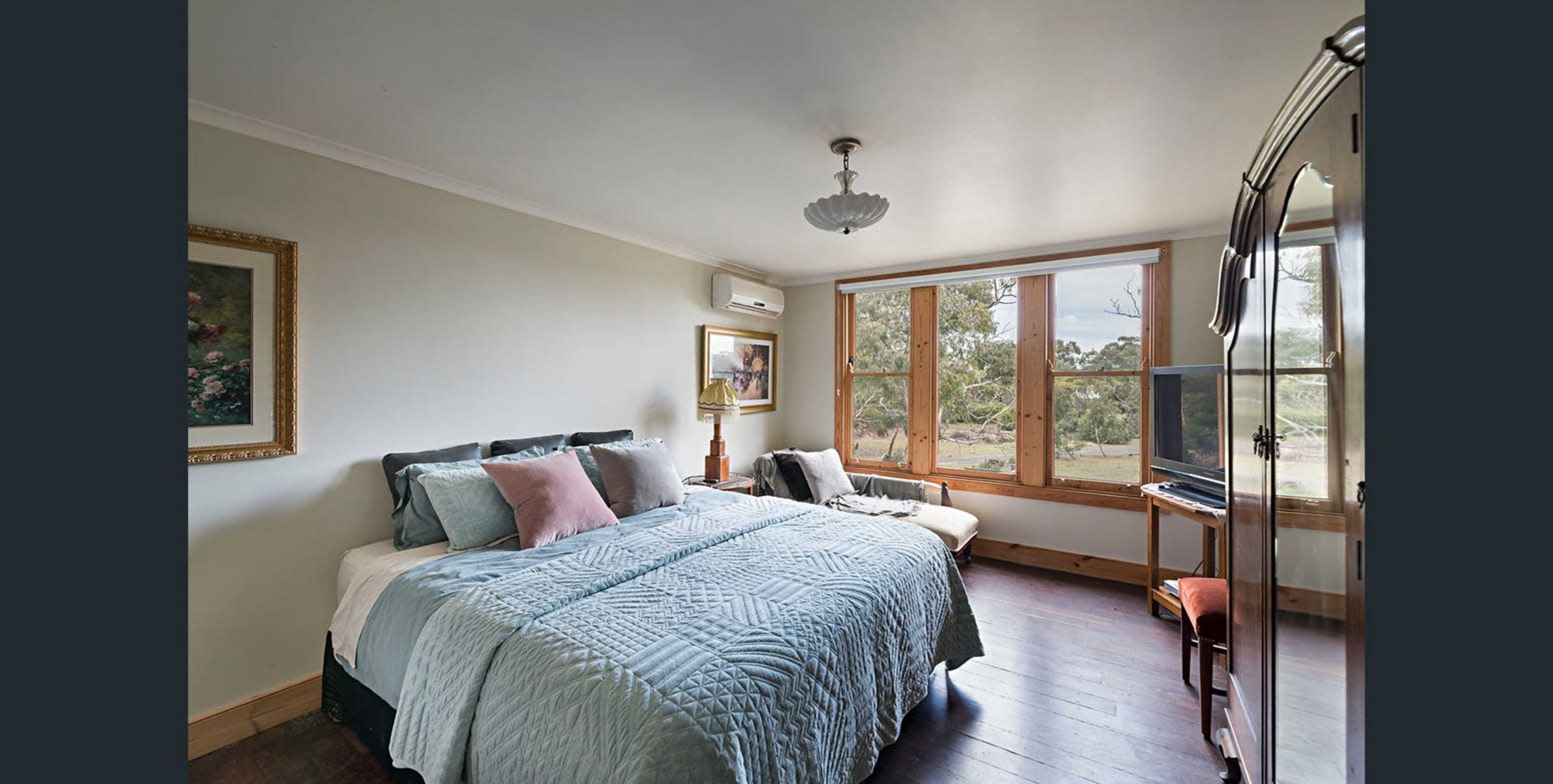 The image shows a well-lit bedroom featuring a bed with a light blue quilt, a nightstand, a large window overlooking a landscape, and a television on a stand.