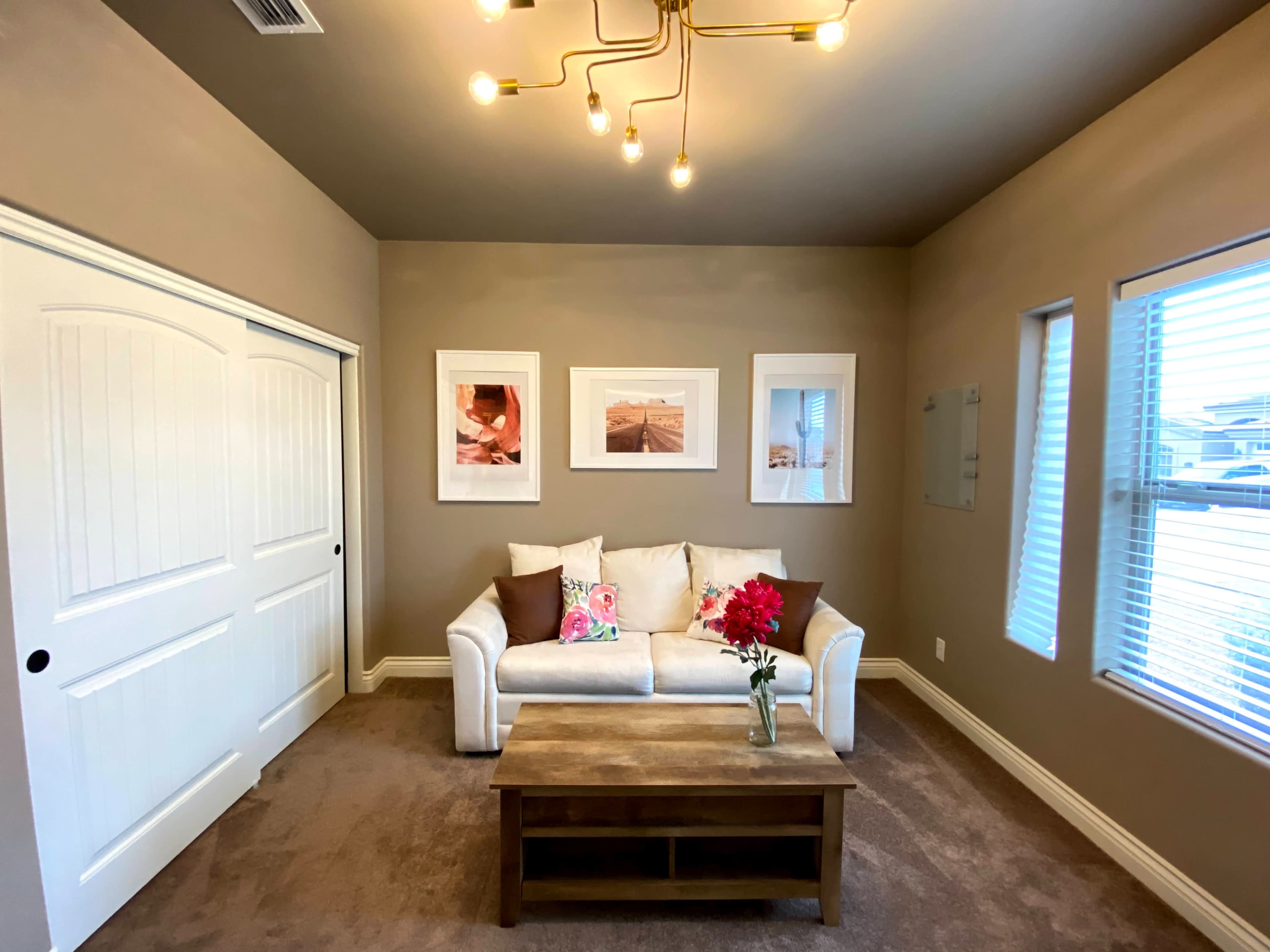 A well-lit living room features a white couch with decorative pillows, a wooden coffee table, and three framed photos on the wall beside large windows.