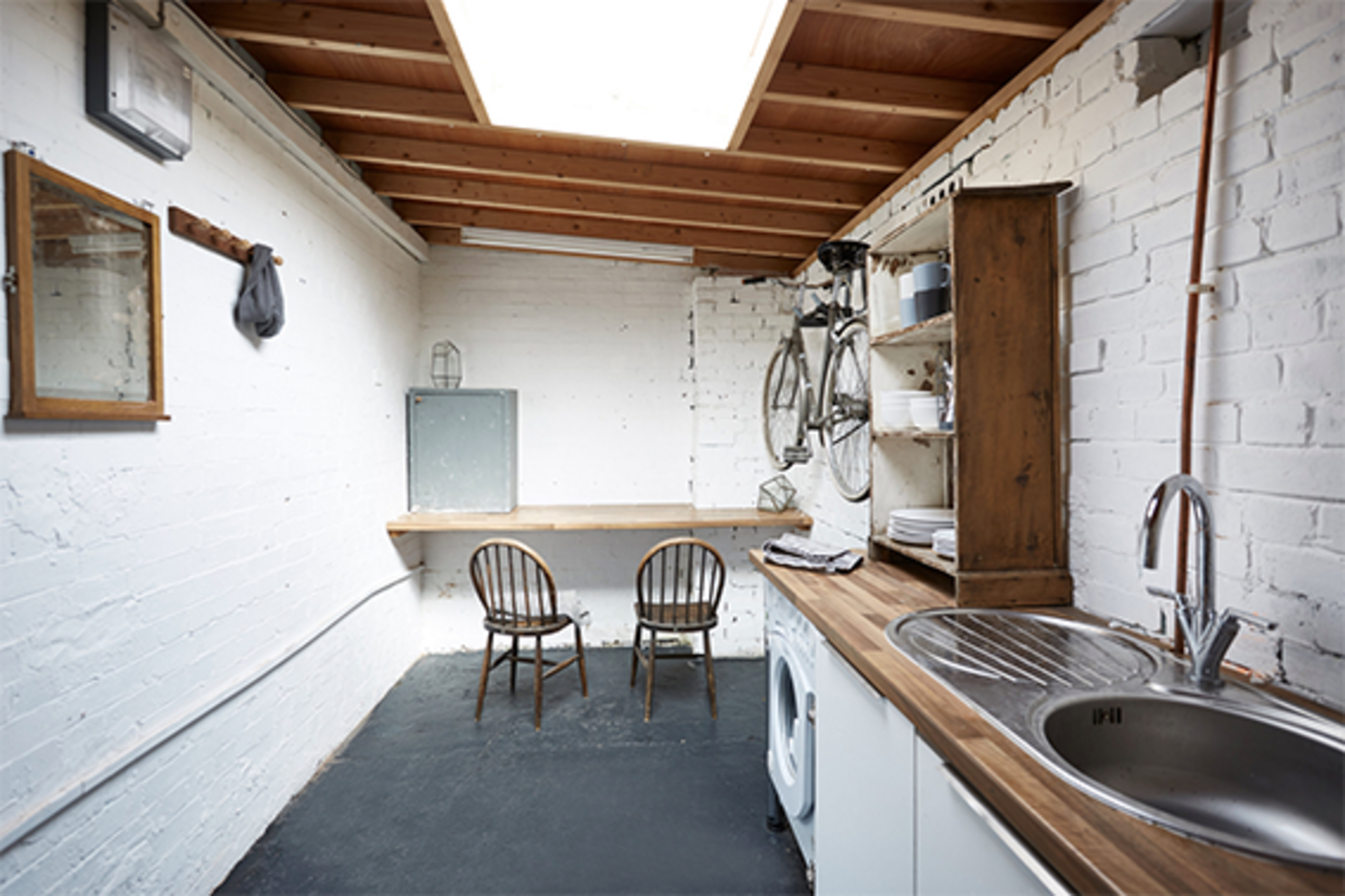 The image shows a minimalist kitchen with white brick walls, a wooden countertop, two wooden chairs, and a washing machine.