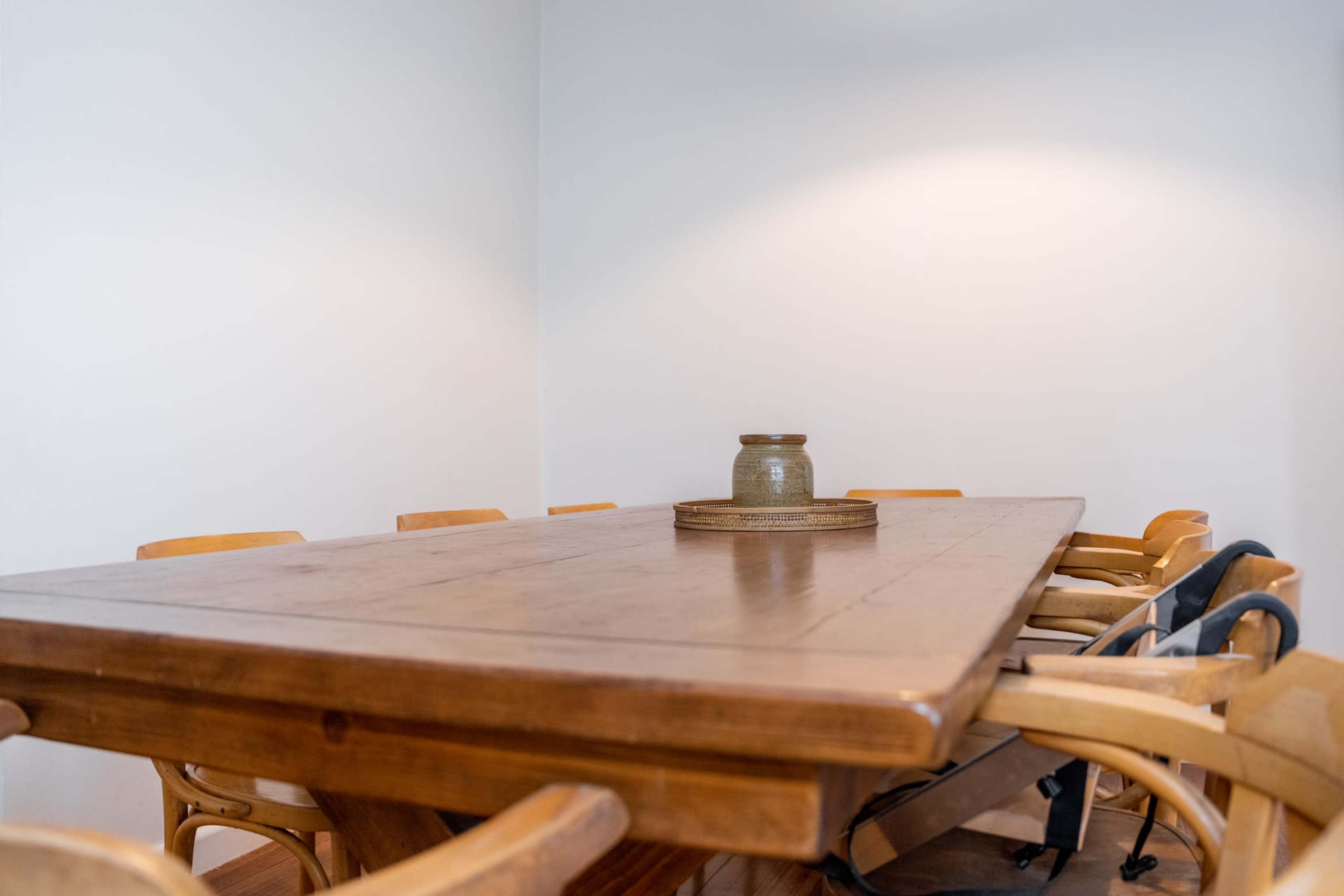 A wooden dining table with a ceramic vase in the center is surrounded by several wooden chairs in a sparsely decorated room.