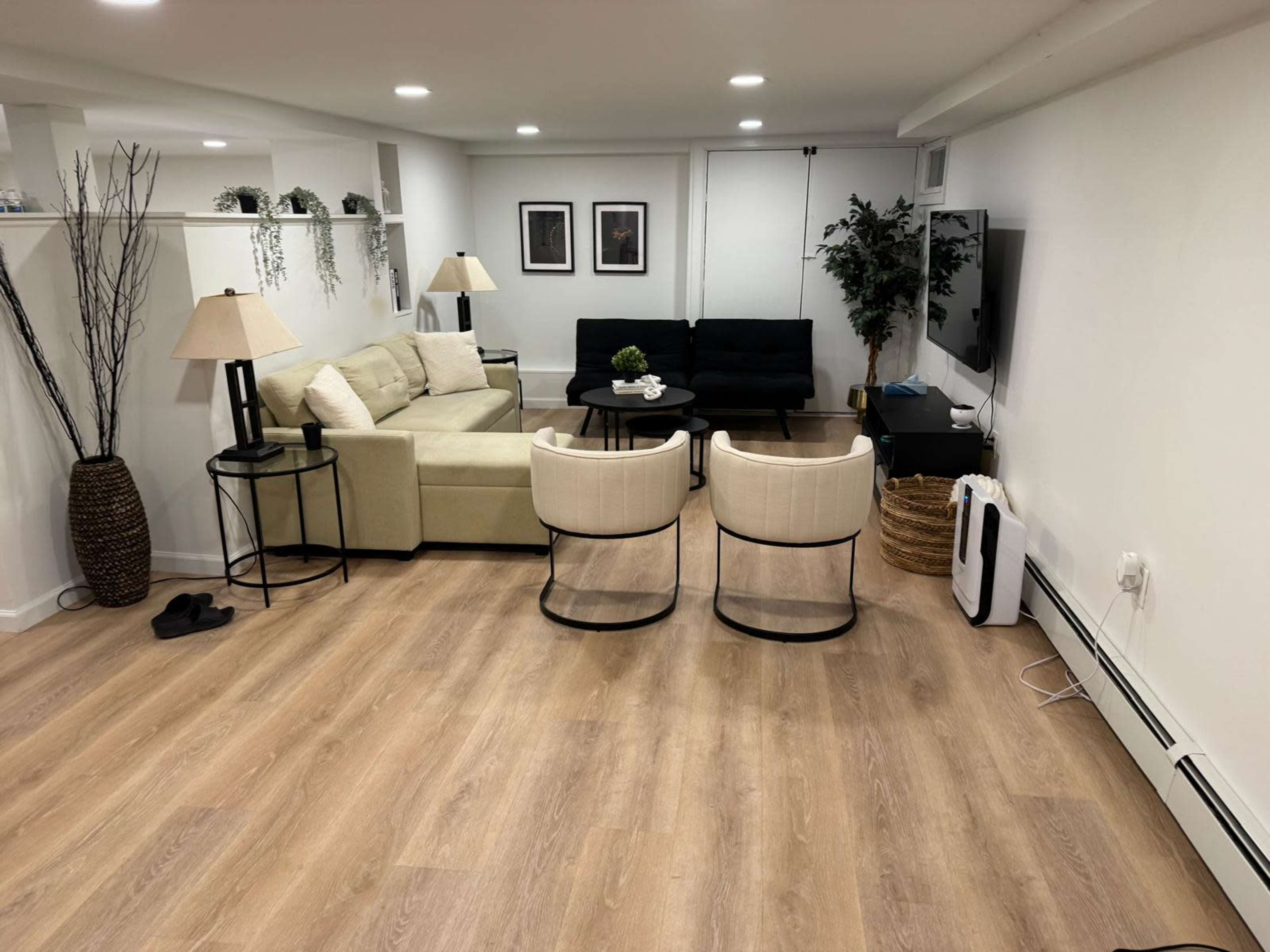 The image shows a finished basement with a light-colored sofa, two armchairs, a television, and decorative plants, all arranged around a wooden floor.