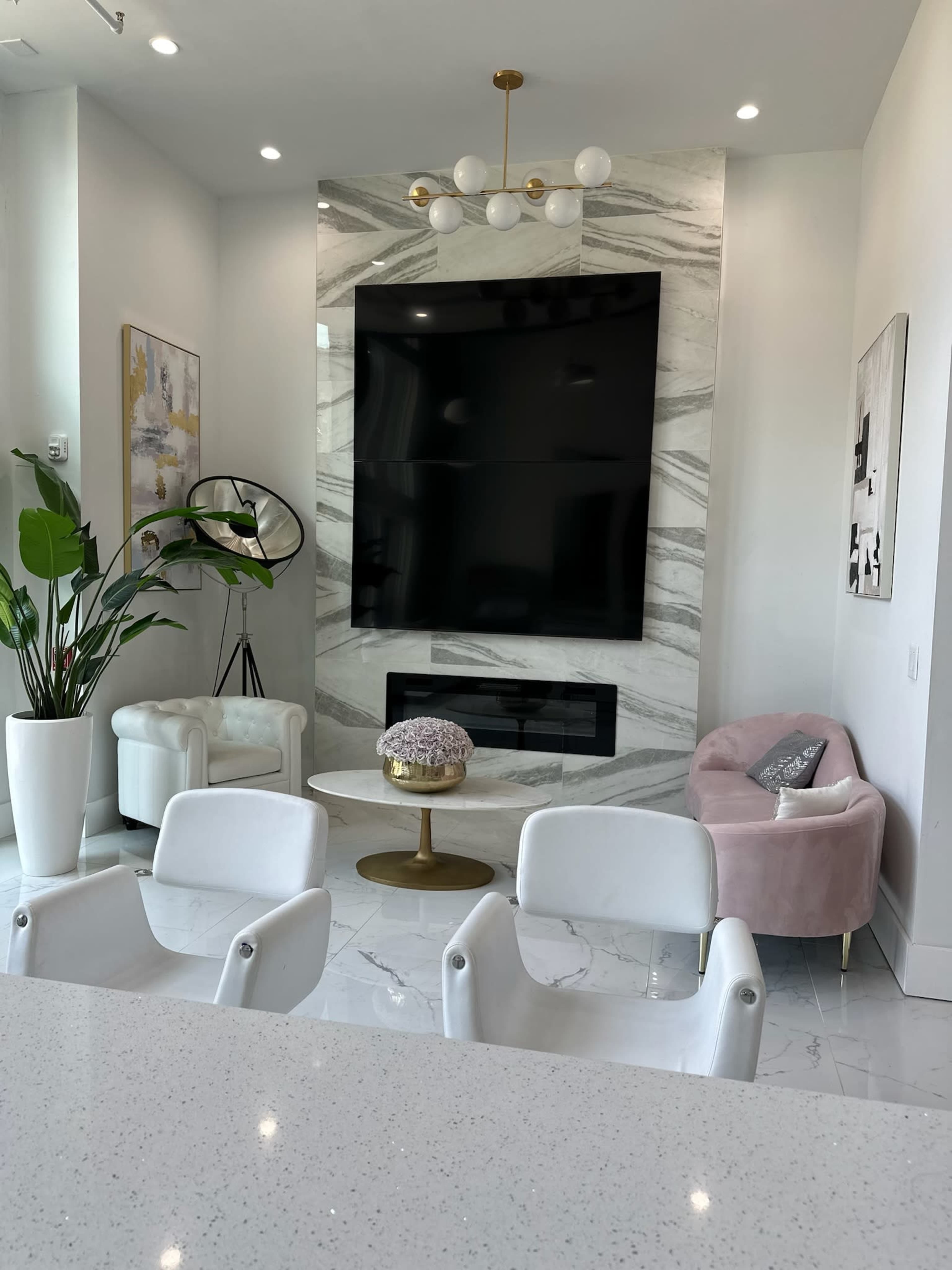 A modern living room features a large black TV mounted on a marble wall, surrounded by white furniture, including a pink chair and a round coffee table.