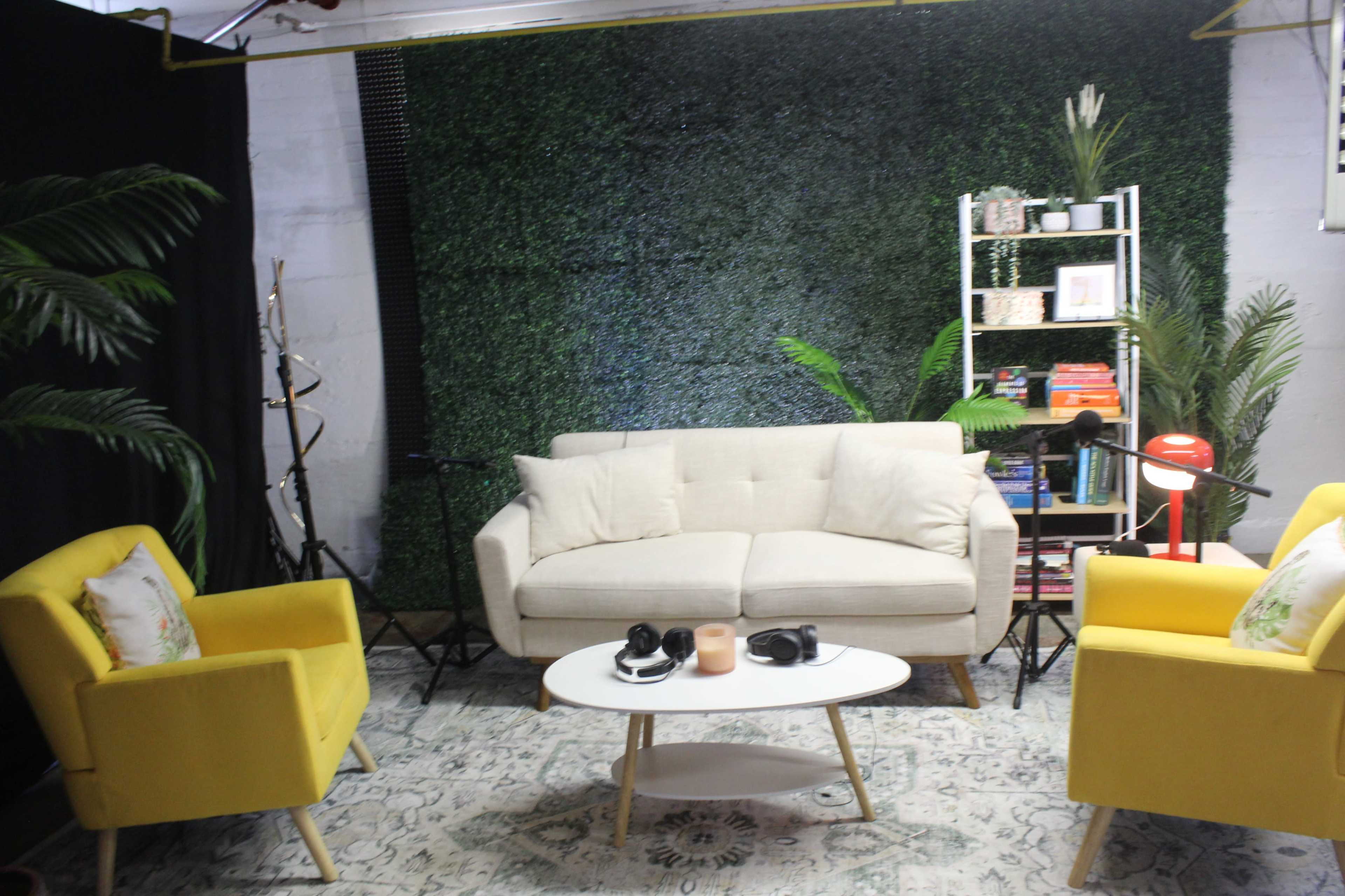 The image shows a cozy studio setup with a cream-colored sofa and two yellow armchairs, surrounded by a green wall, a small white coffee table, and various decorative items.