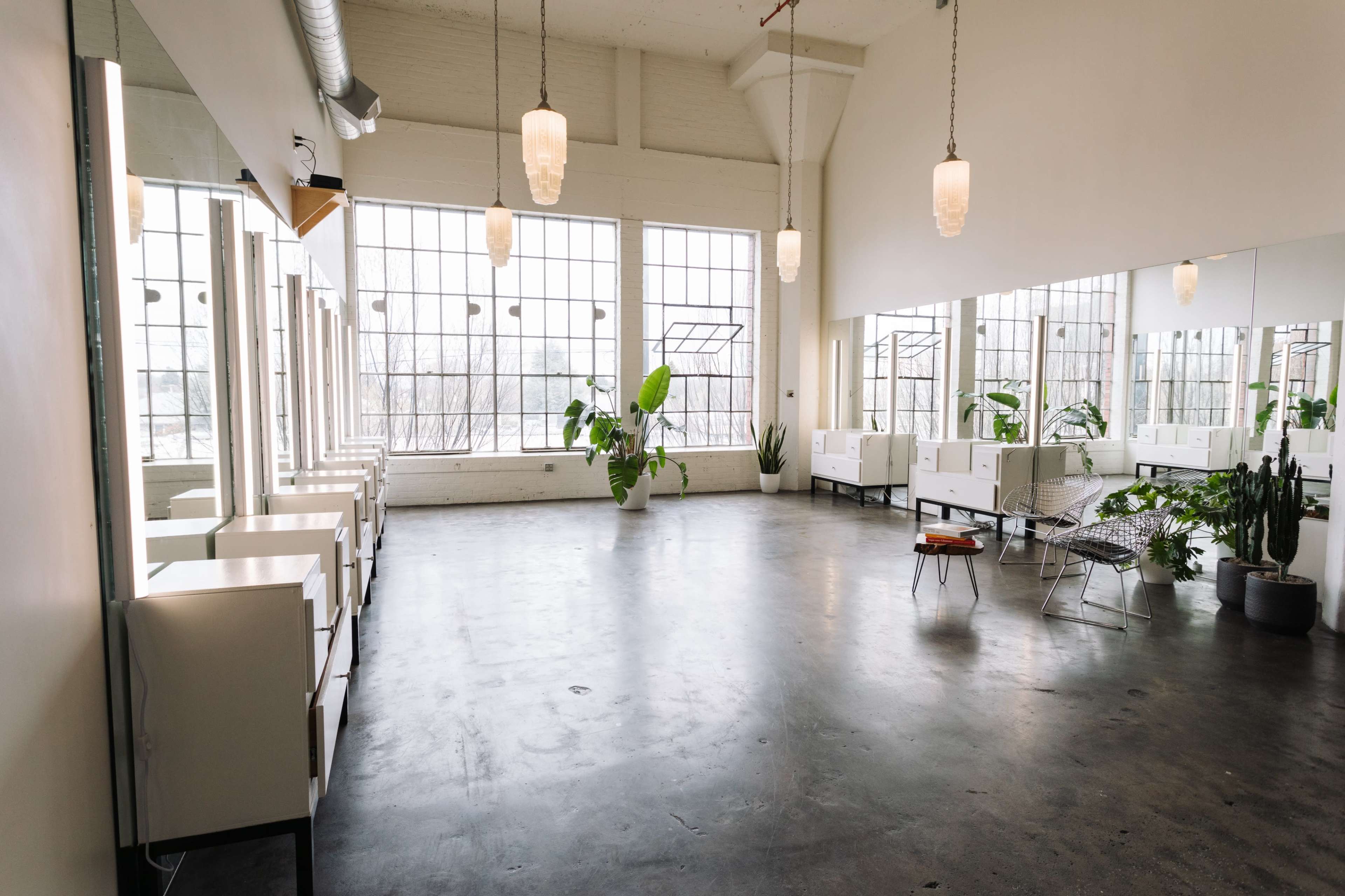 A minimalist salon interior with large windows, several hair styling stations, and various indoor plants.