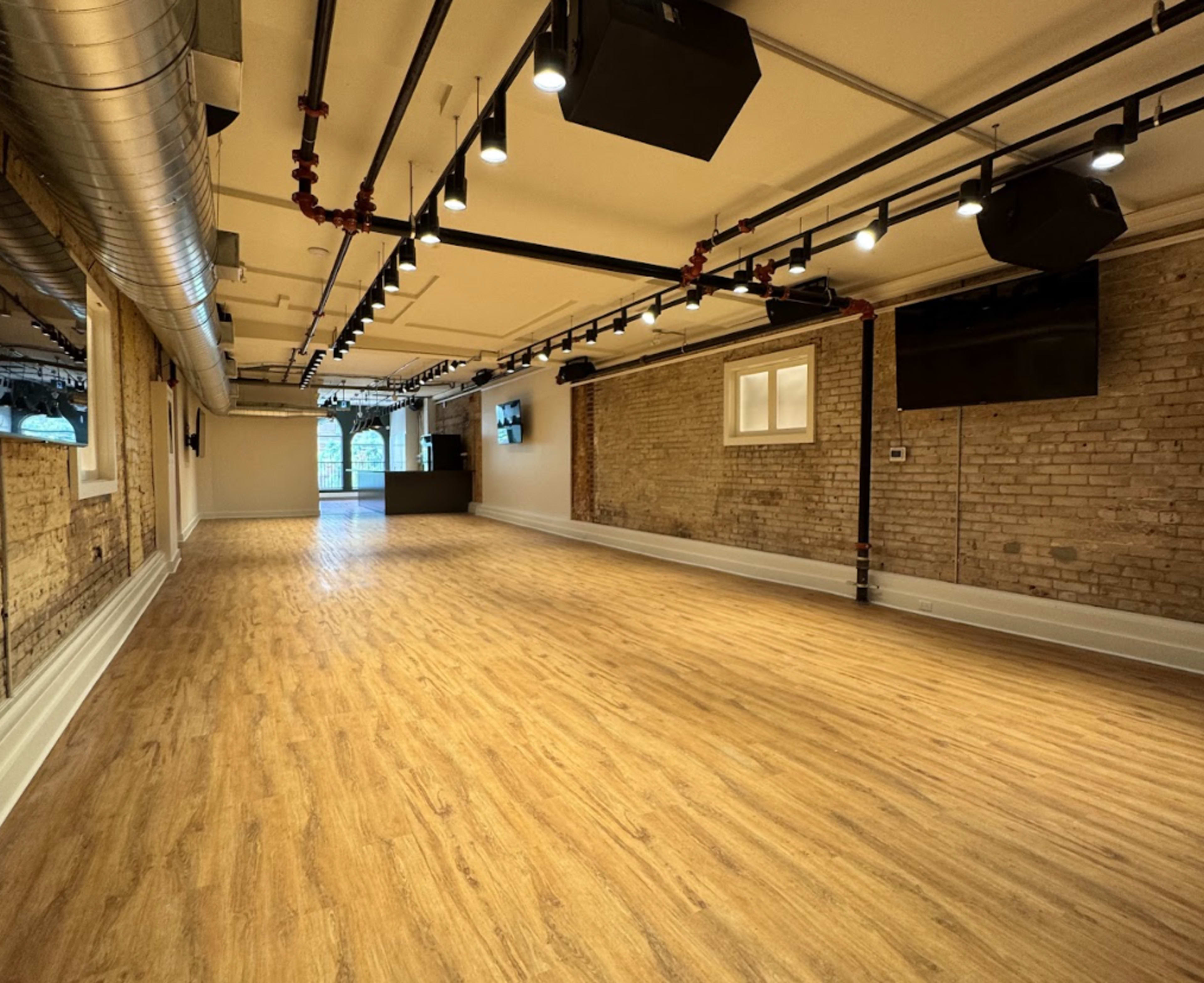 The image shows a spacious room with wooden flooring, exposed brick walls, and various ceiling lights.