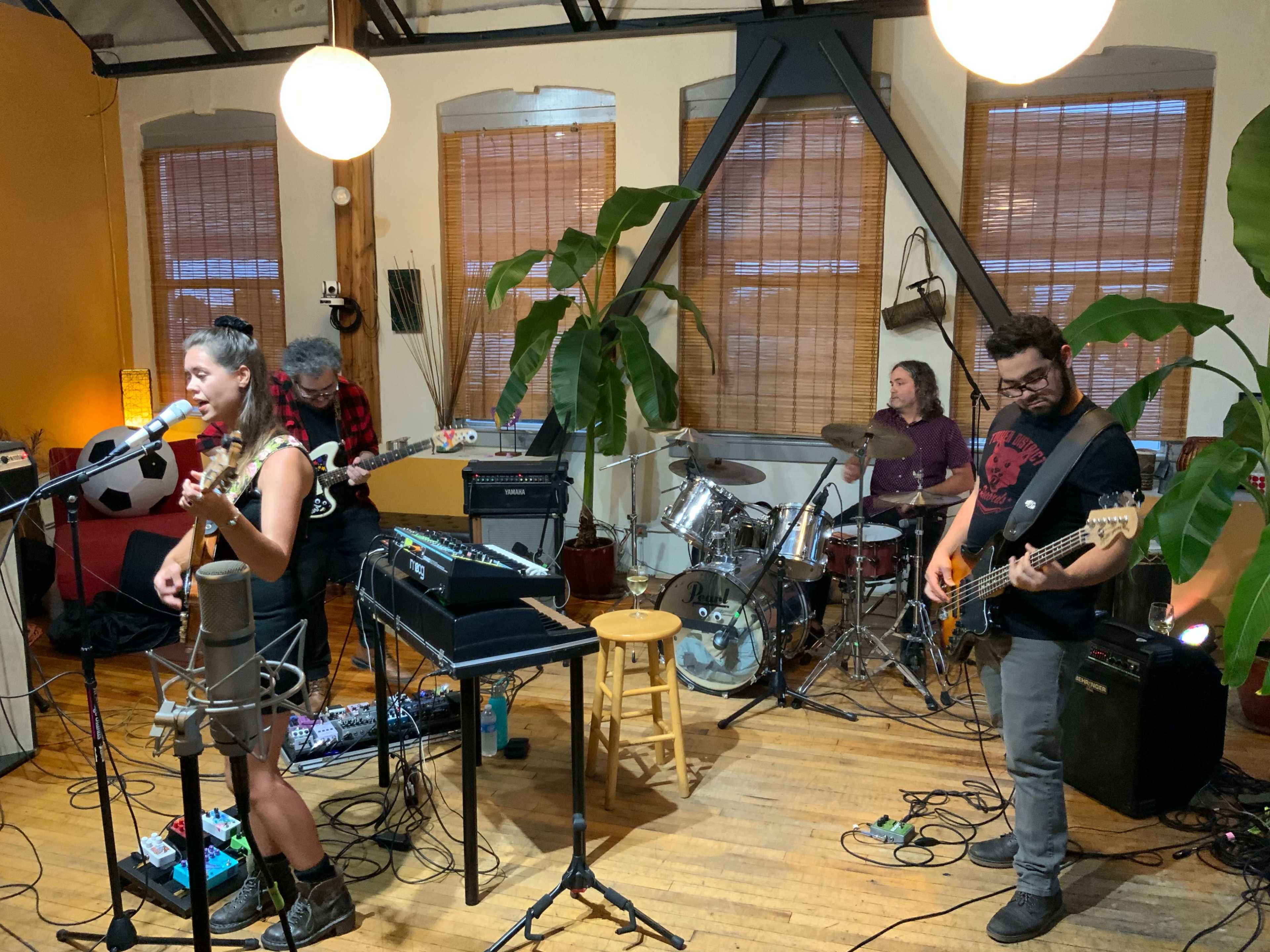 A band performs in a cozy indoor space with musical instruments and plants arranged around them.