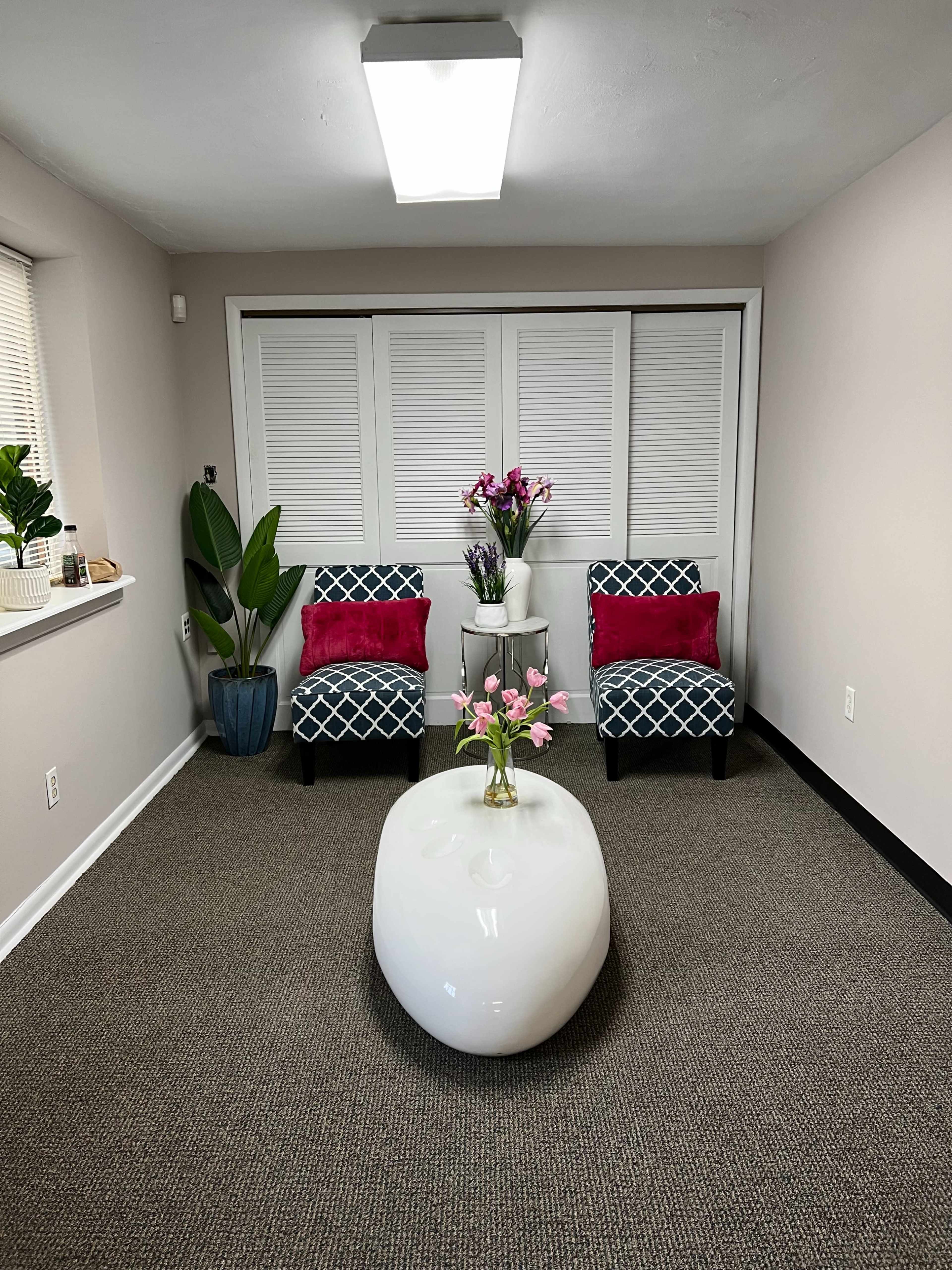The image shows a well-lit room with two patterned chairs, a round white coffee table, and decorative flowers in a vase, accompanied by potted plants.