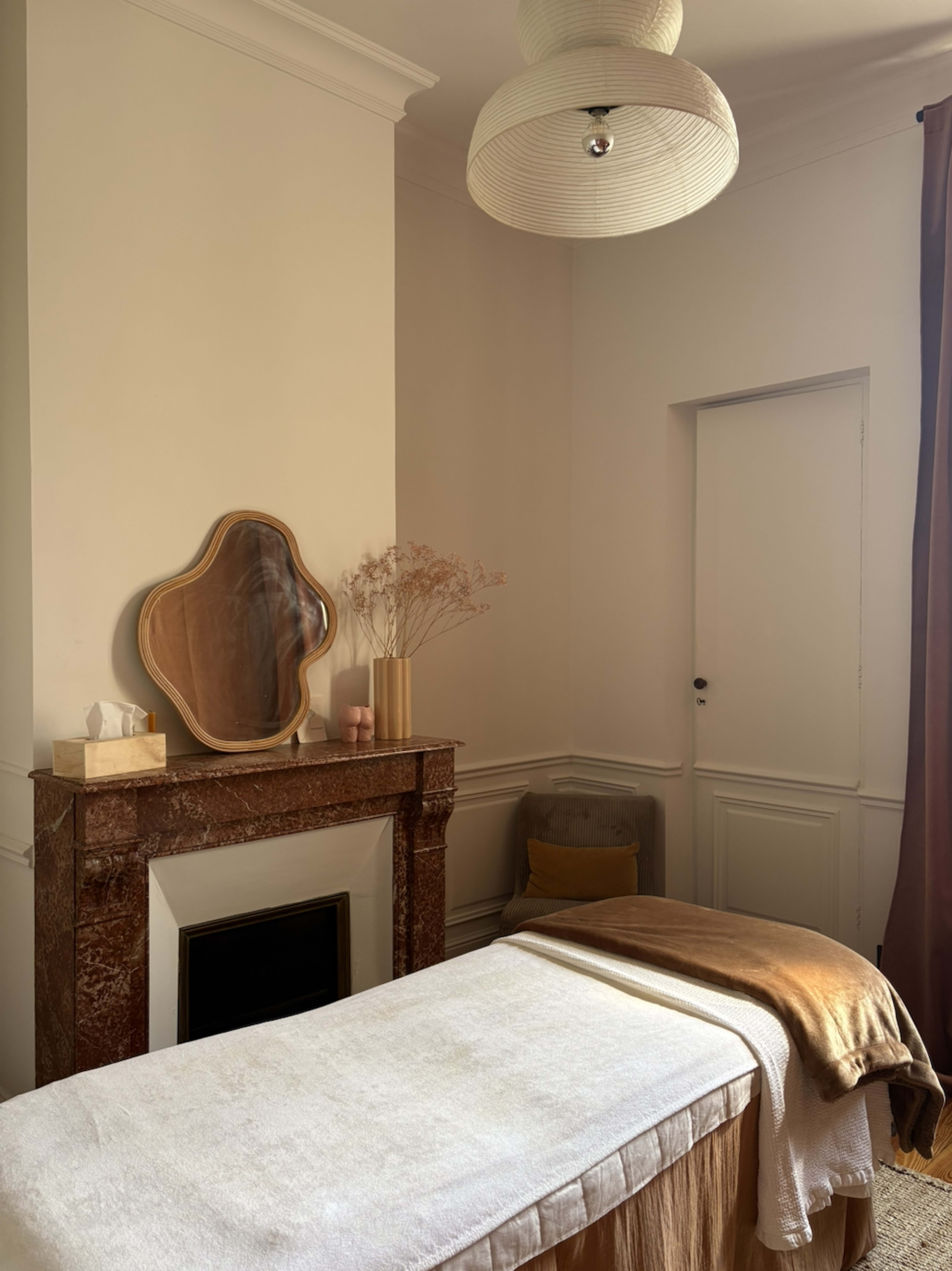 The image shows a tranquil room featuring a massage table, a decorative fireplace with a mirror above it, and soft furnishings in warm tones.