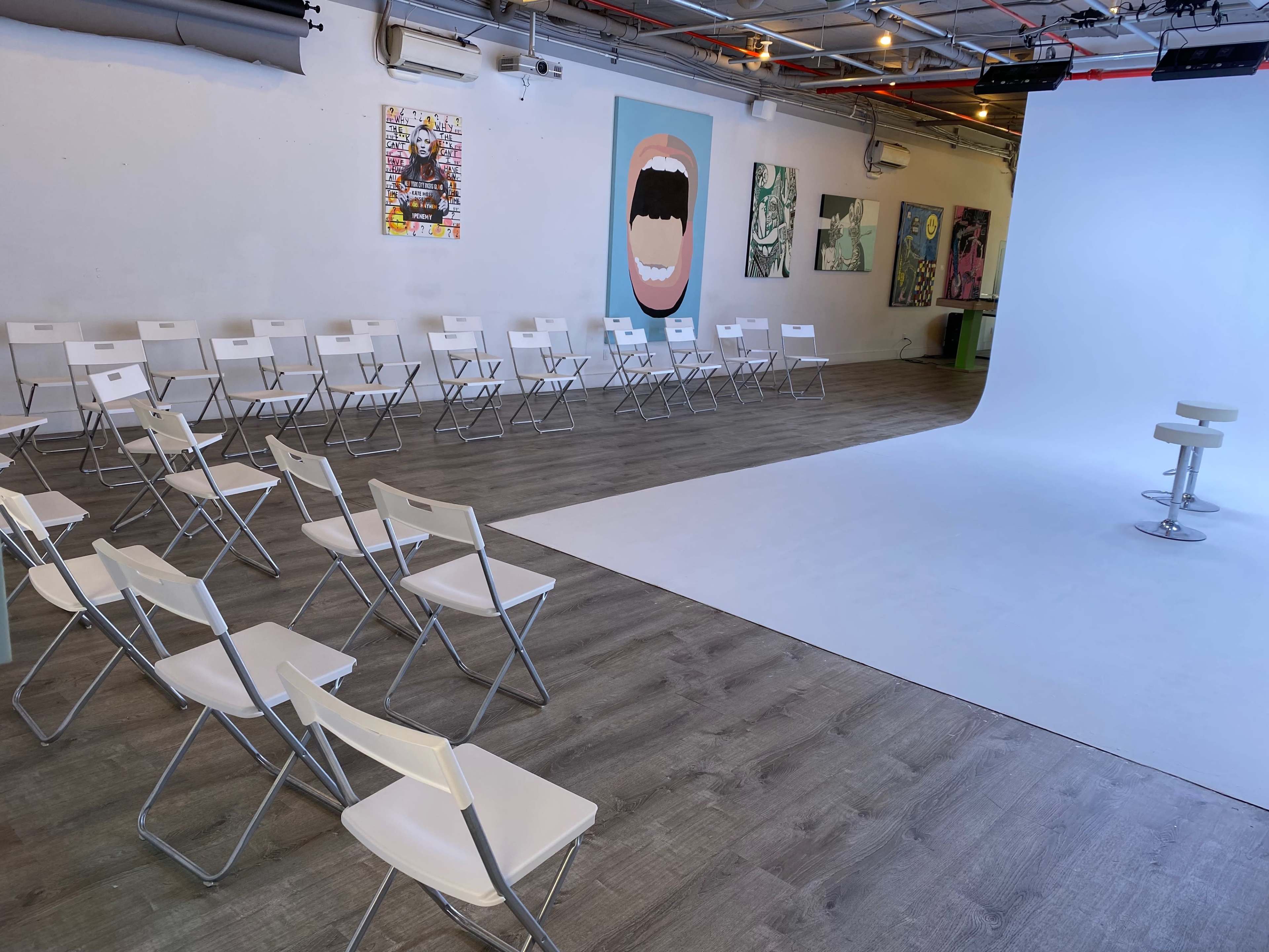 The image shows a spacious art studio with white folding chairs arranged in rows facing a white backdrop, flanked by colorful artworks on the walls.