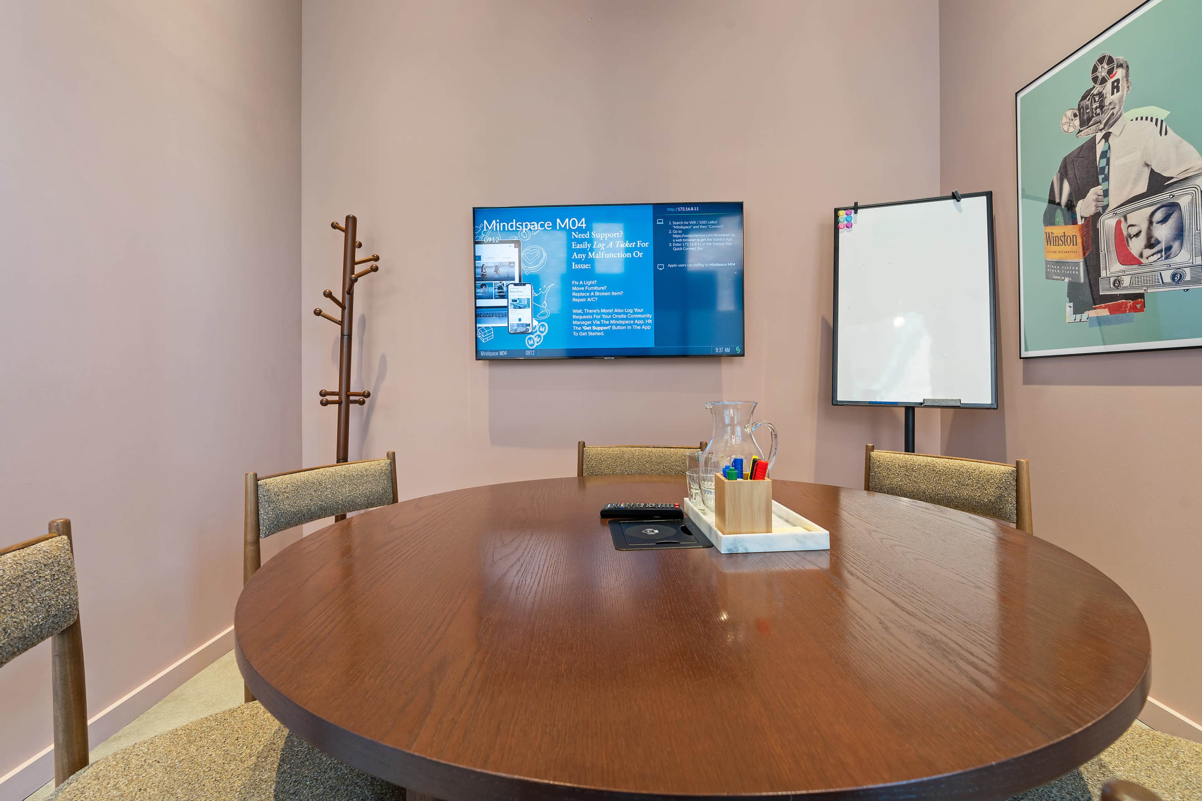 A conference room features a round wooden table, a flat-screen TV displaying content, a whiteboard, and a coat rack against a pink wall.