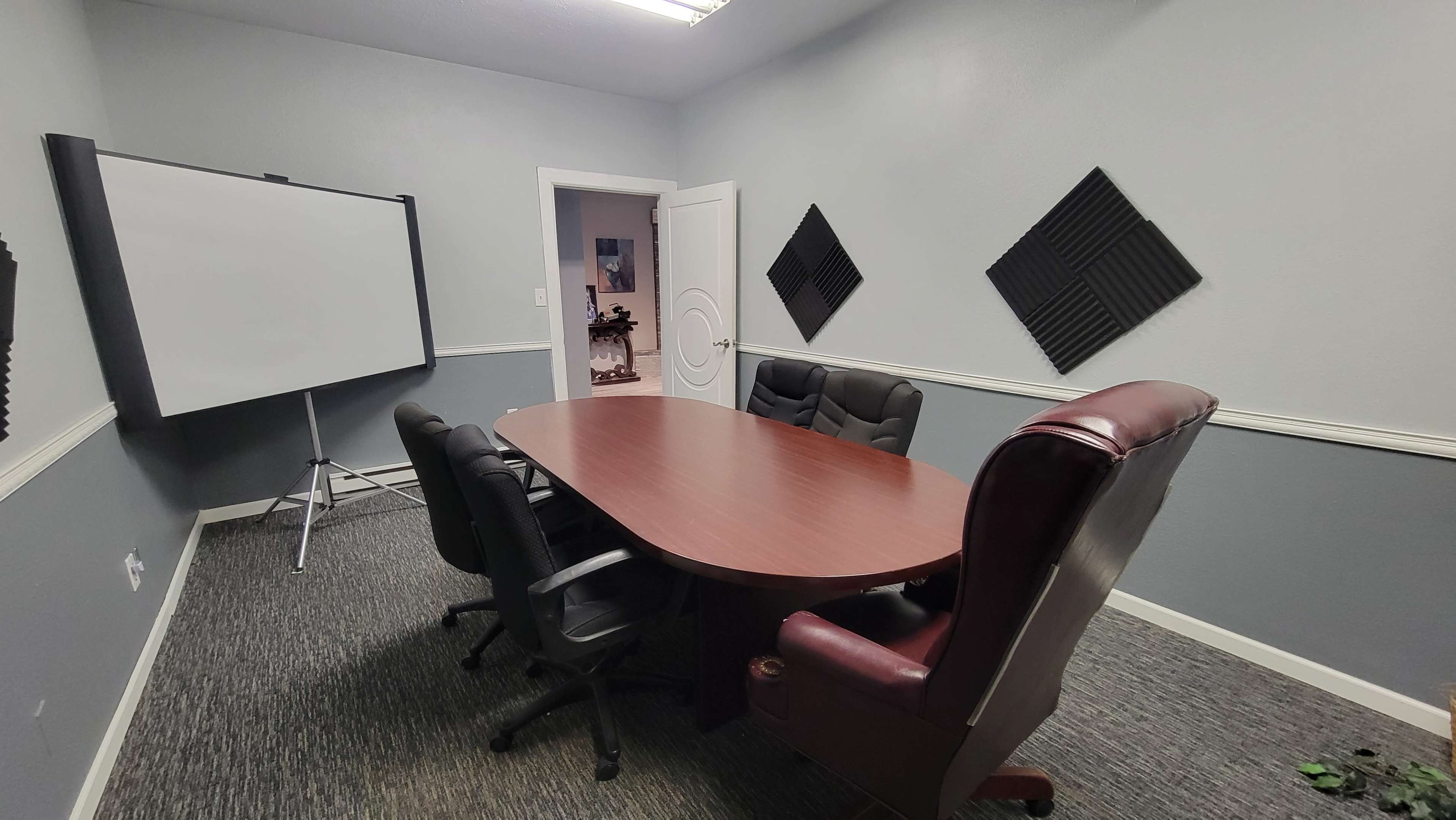 A conference room with a large oval wooden table surrounded by five black chairs and a presentation board on a stand against the wall.