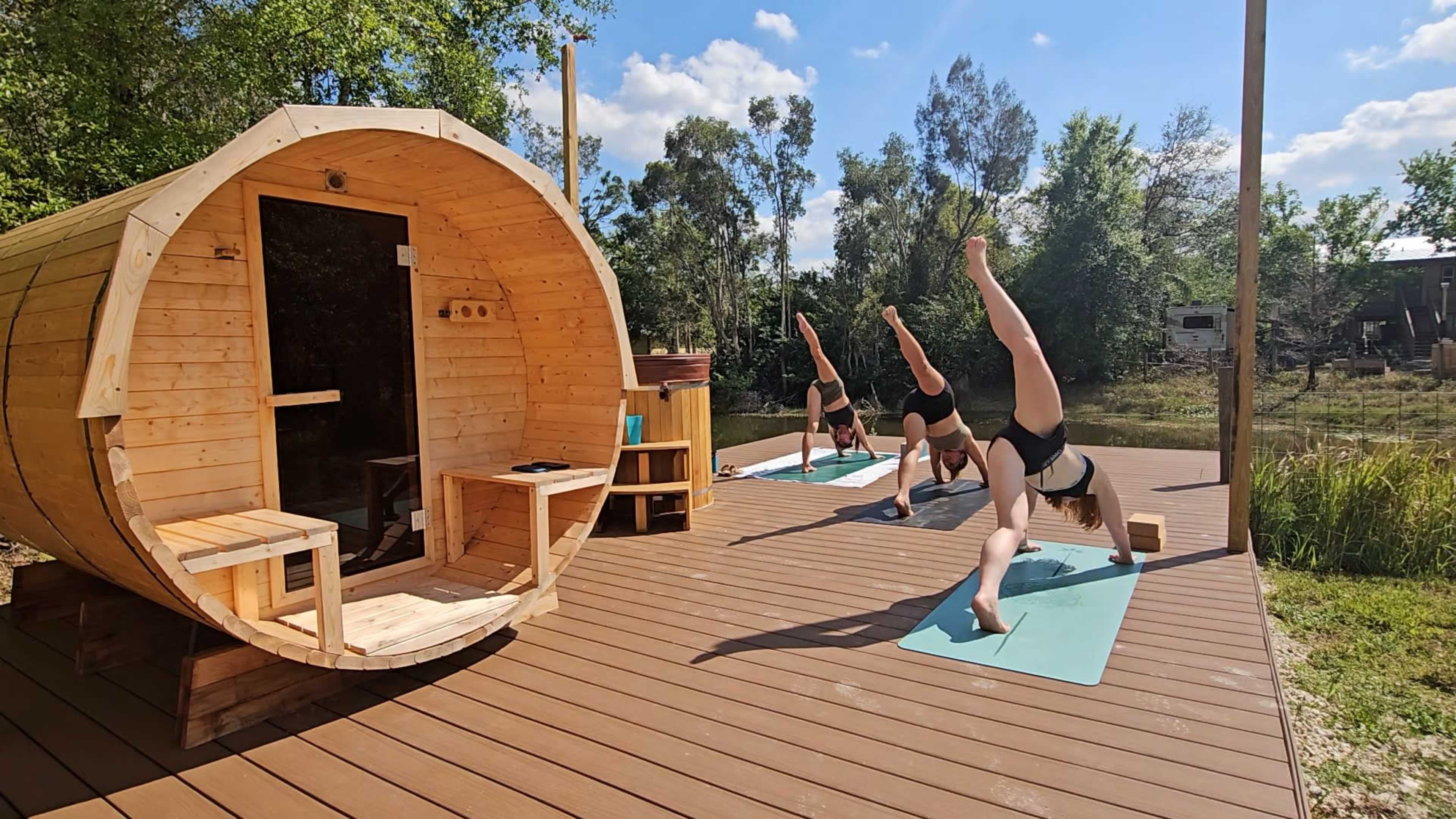 Three individuals perform yoga poses on mats outside a wooden structure surrounded by trees and a pond.