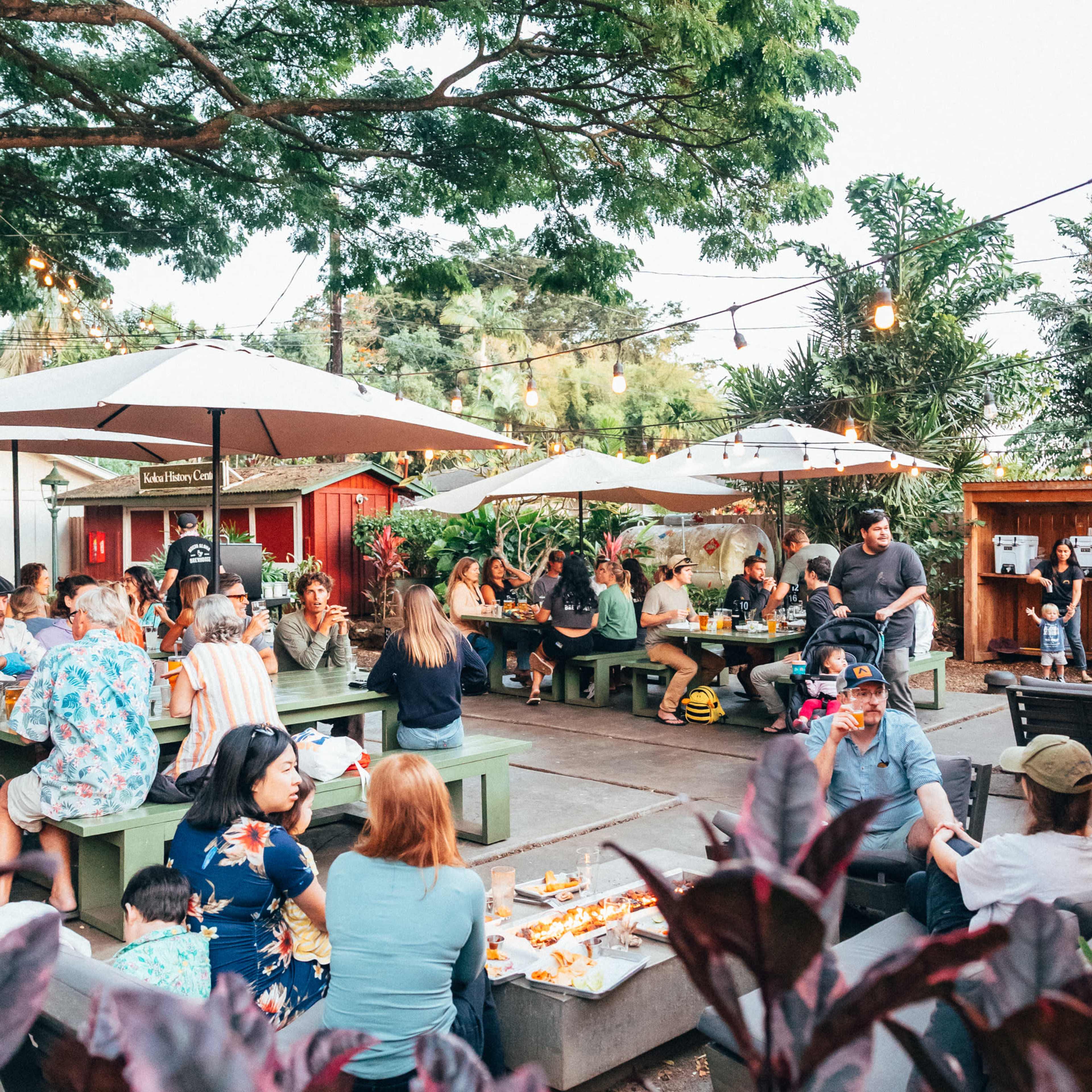 A lively outdoor dining area is filled with people sitting at tables under umbrellas, surrounded by greenery and string lights.