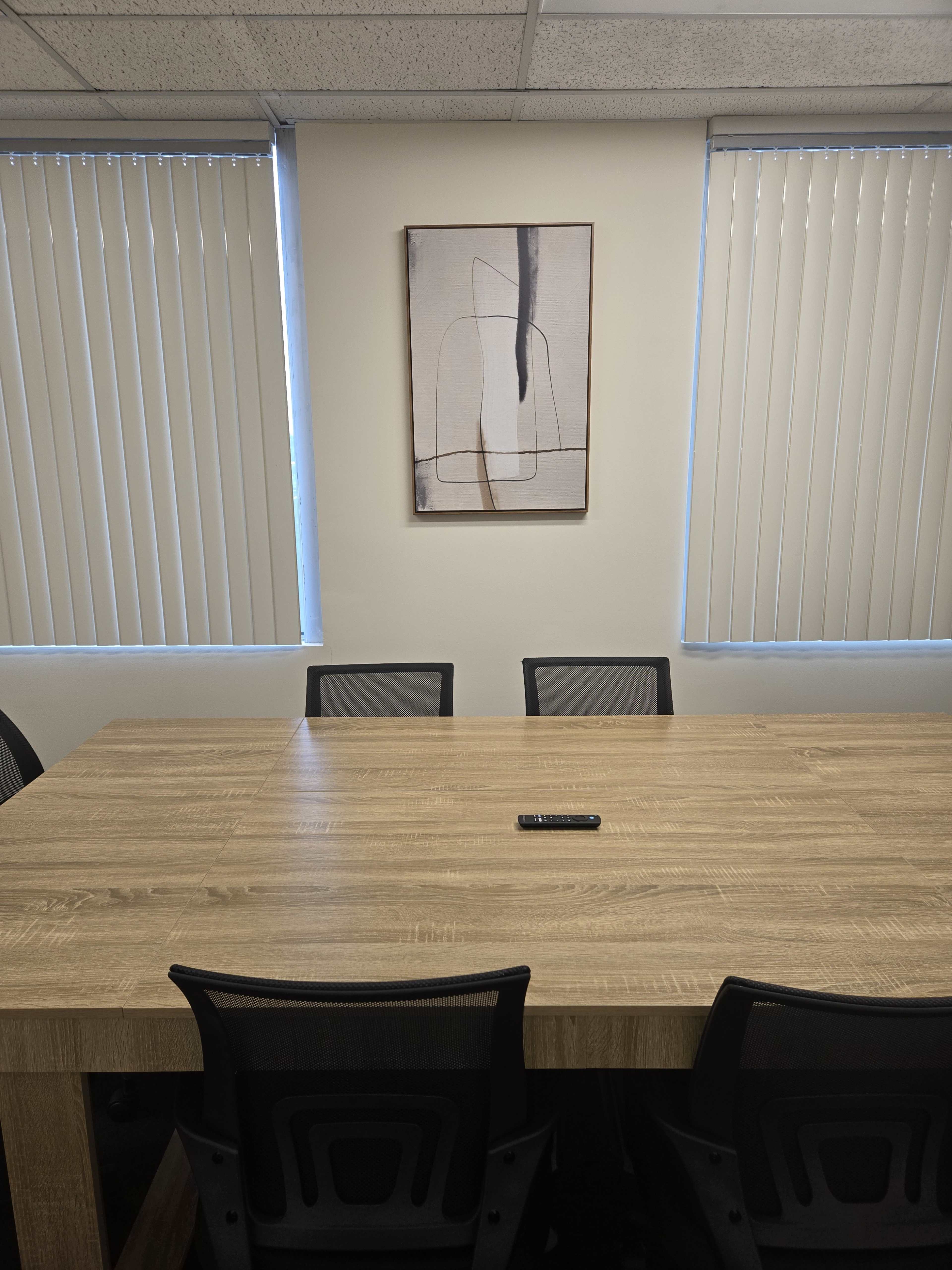 A conference room features a large wooden table surrounded by black mesh chairs, with a work of art hanging on the wall and blinds covering two windows.