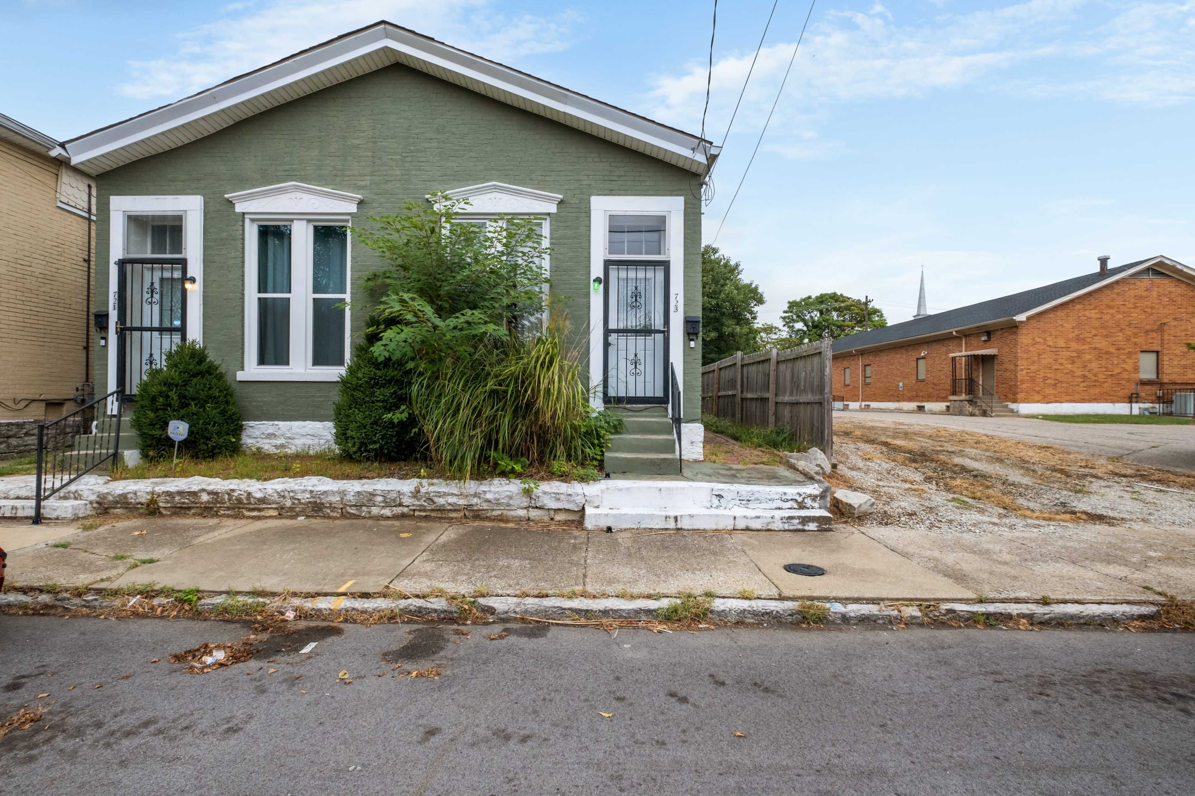 A green house with white trim, featuring two entry doors, surrounded by overgrown plants, located on a residential street.