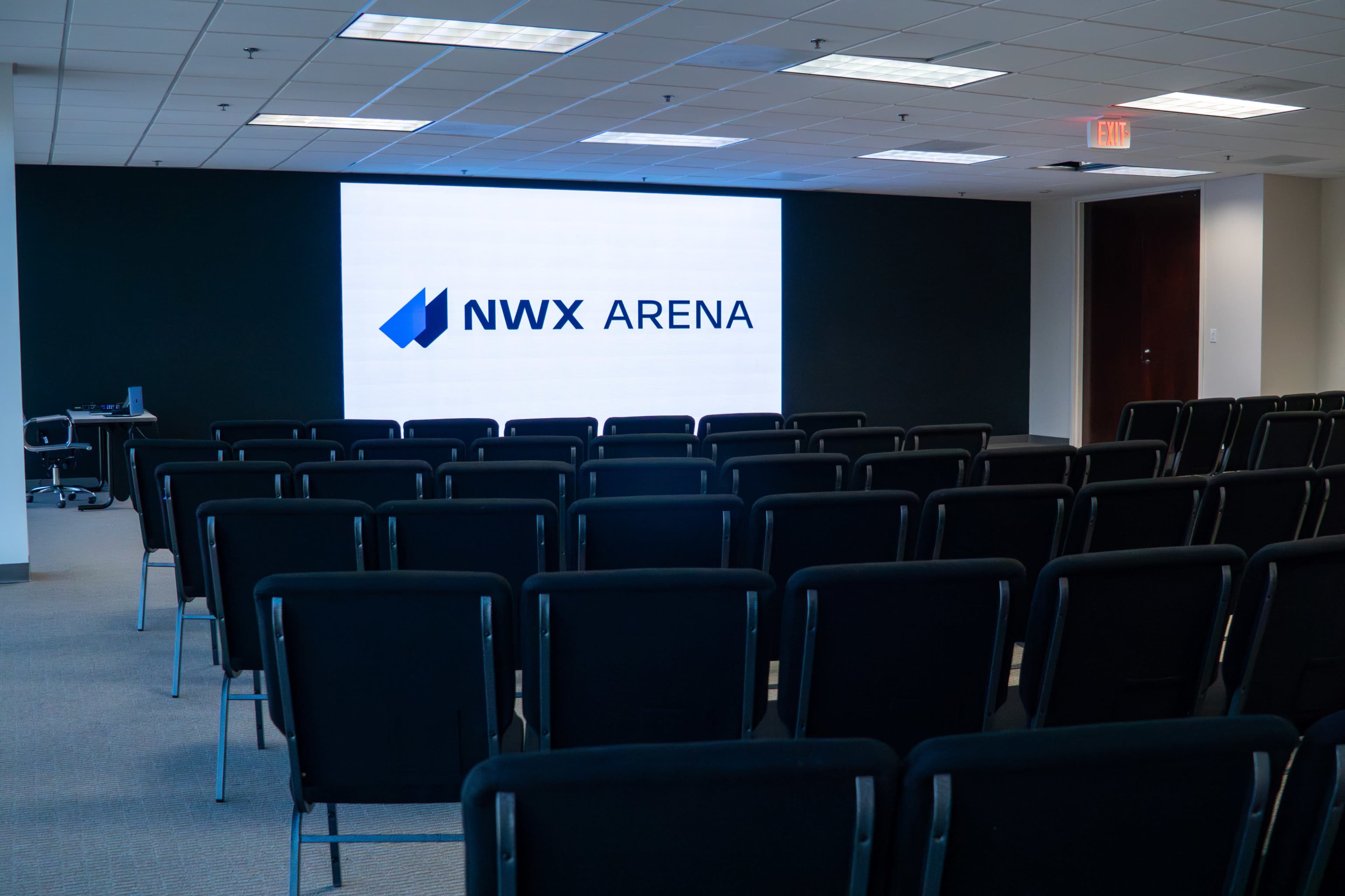 The image shows a seminar room with rows of black chairs facing a large screen displaying the text "NWX ARENA."