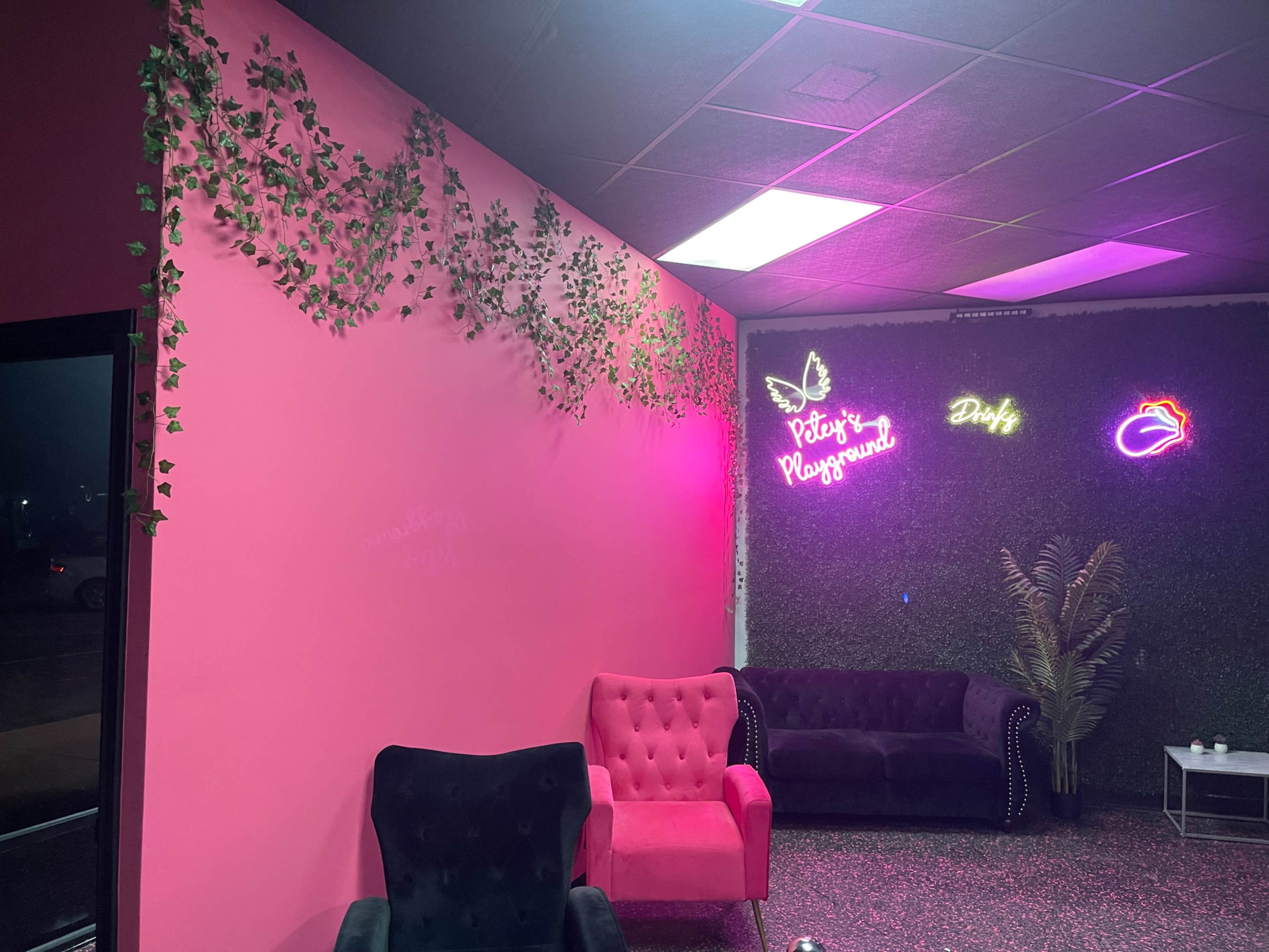 The scene features a brightly colored pink wall adorned with green vines, against which neon signs and plush seating are arranged in a stylish interior.