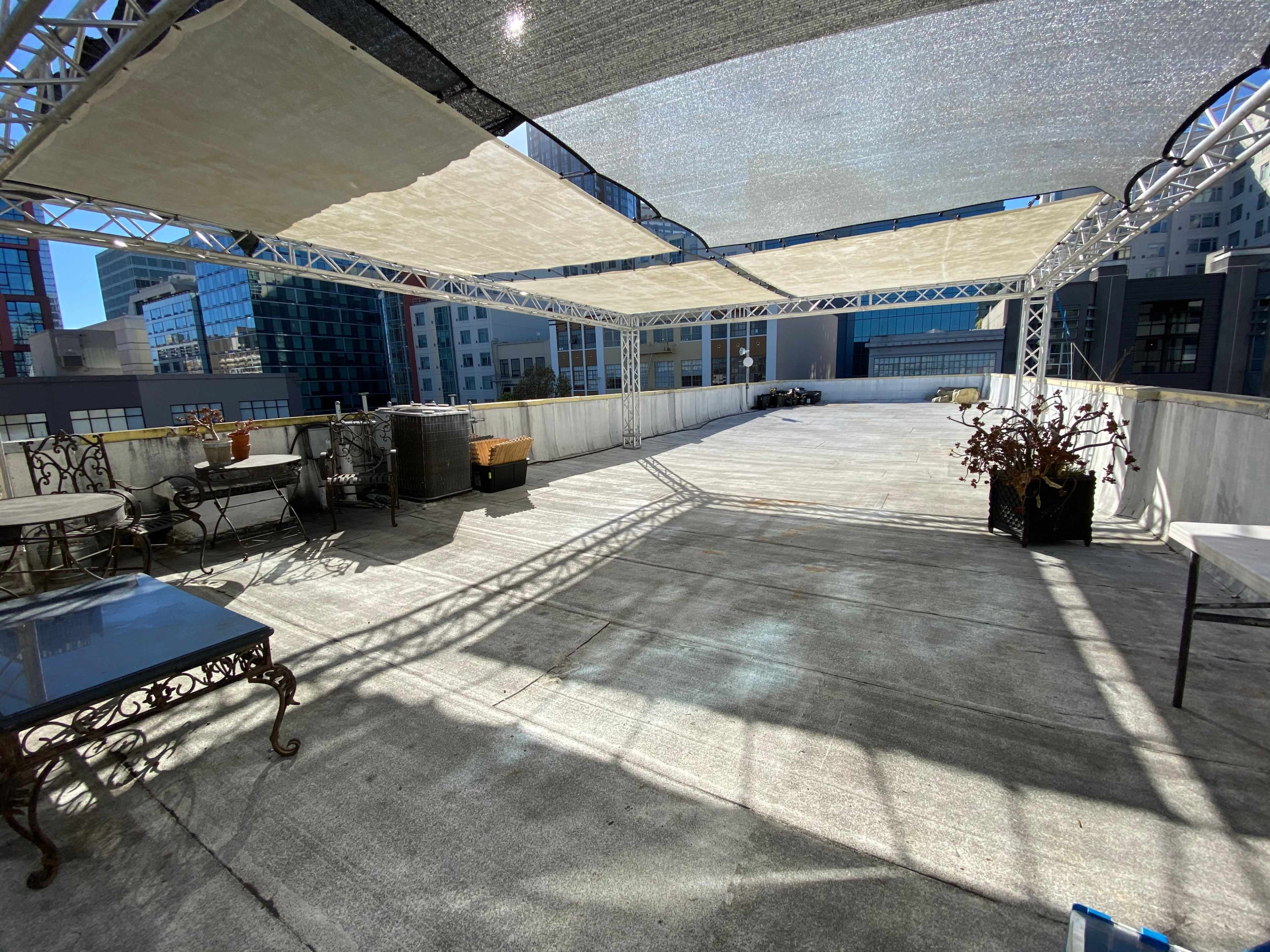 A rooftop area with a clear concrete surface, featuring metal-framed tables and chairs, shaded by large fabric canopies.