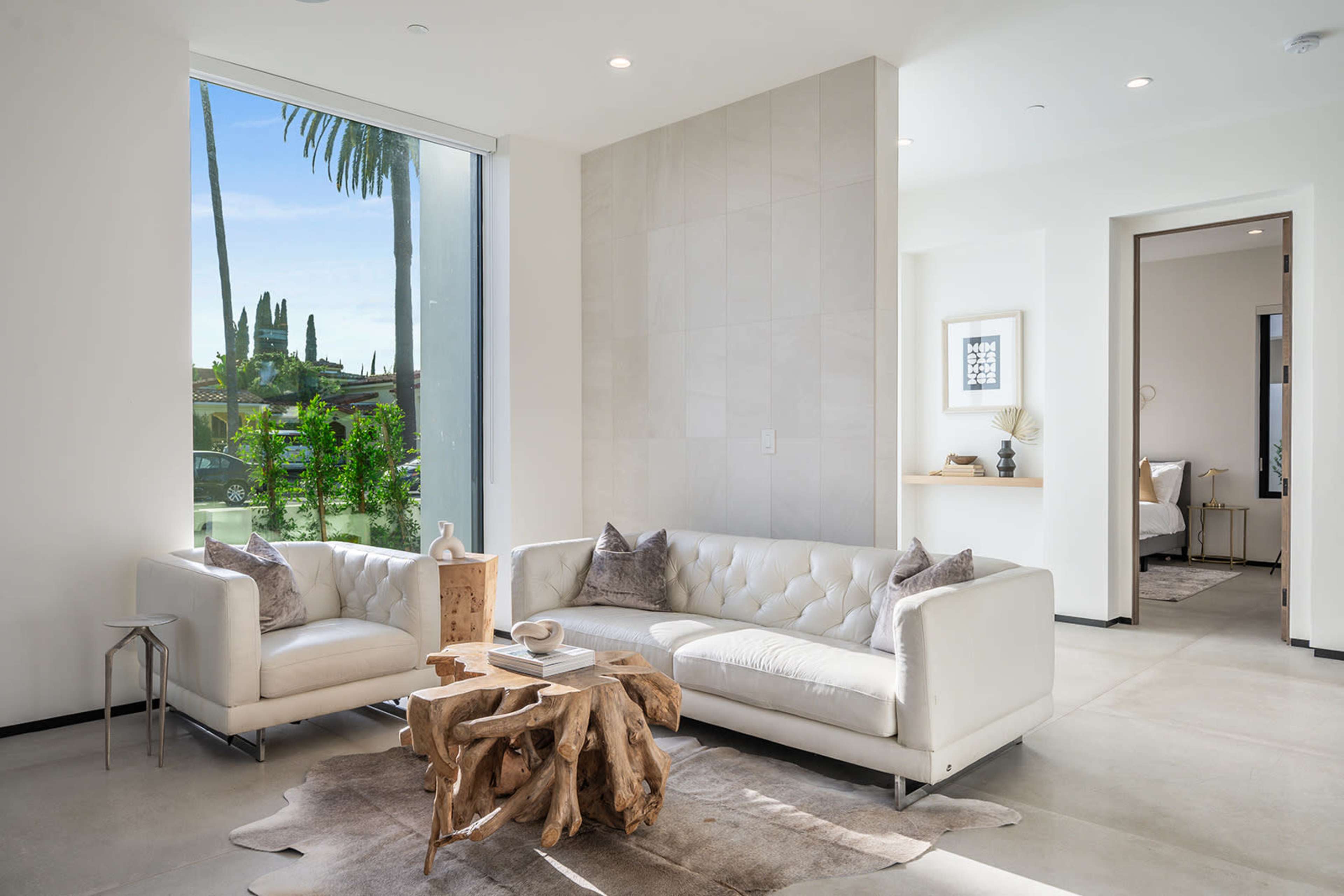 A modern living room features a white sectional sofa, a unique wooden coffee table, and large windows allowing natural light to flow in.