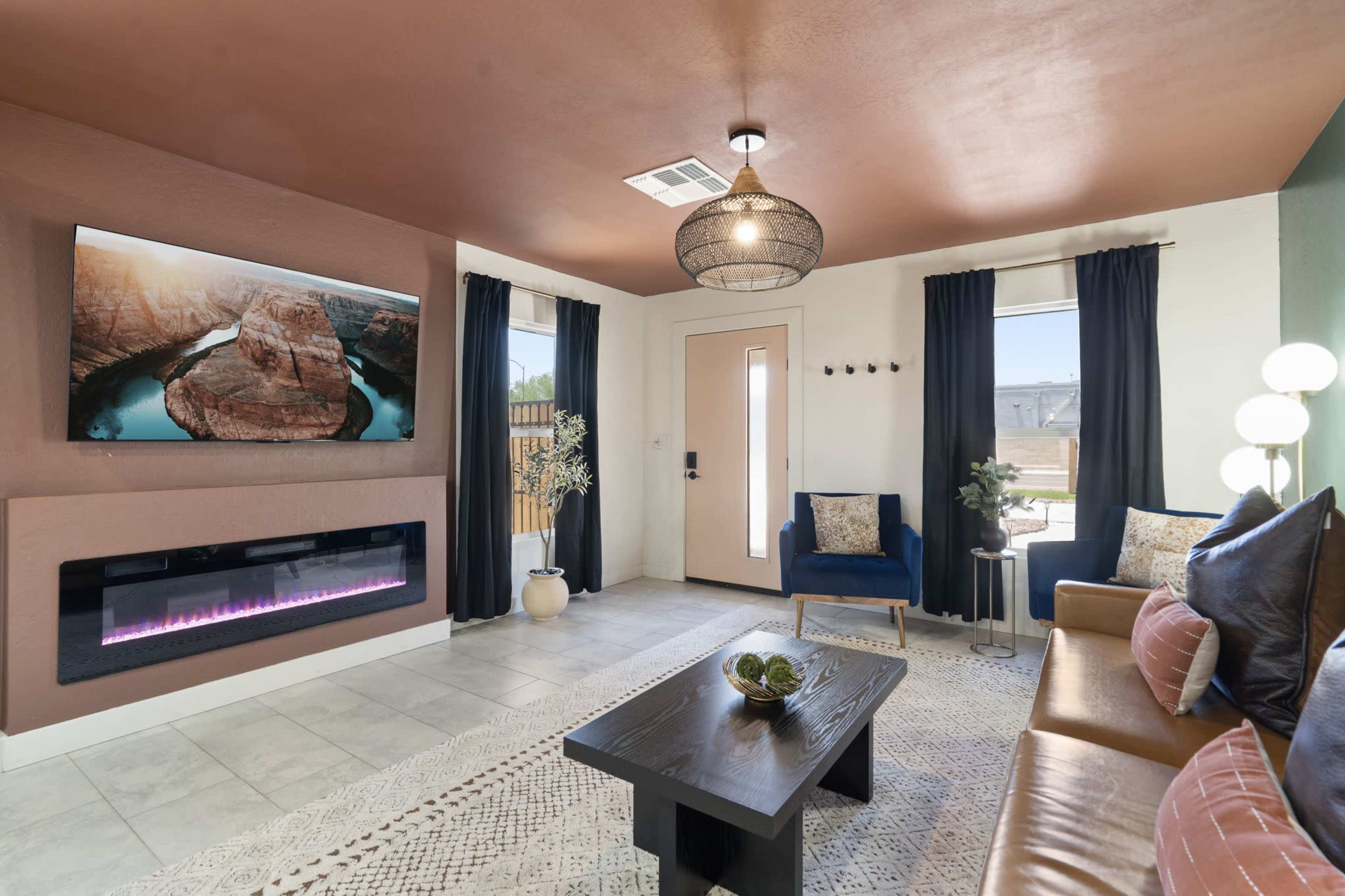 The image shows a modern living room featuring a mounted television, an electric fireplace, two blue chairs, a brown sofa, and large windows with sheer curtains.