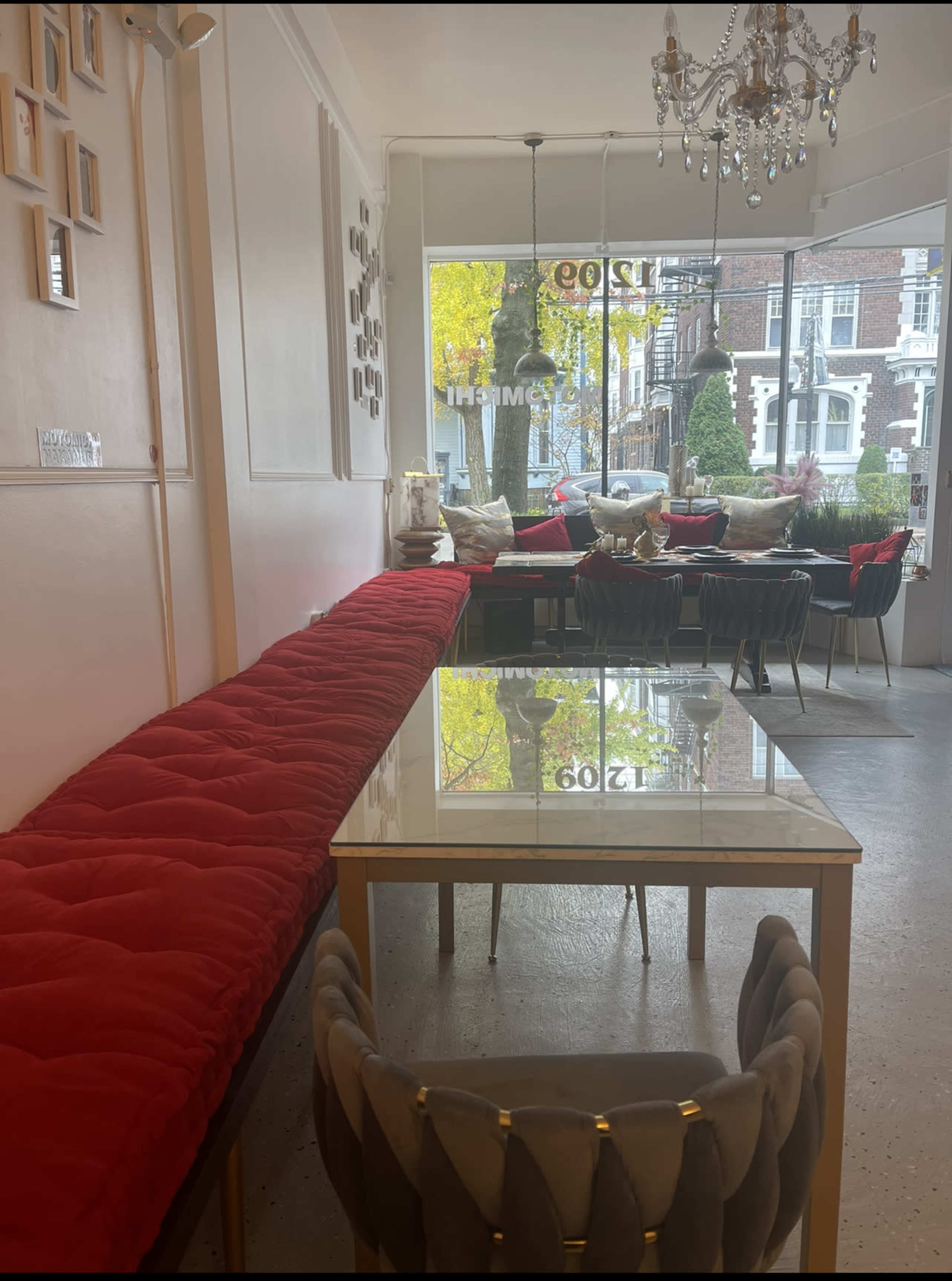 A spacious room features a long red upholstered bench, a glass-topped table, and a chandelier, with large windows showcasing greenery outside.