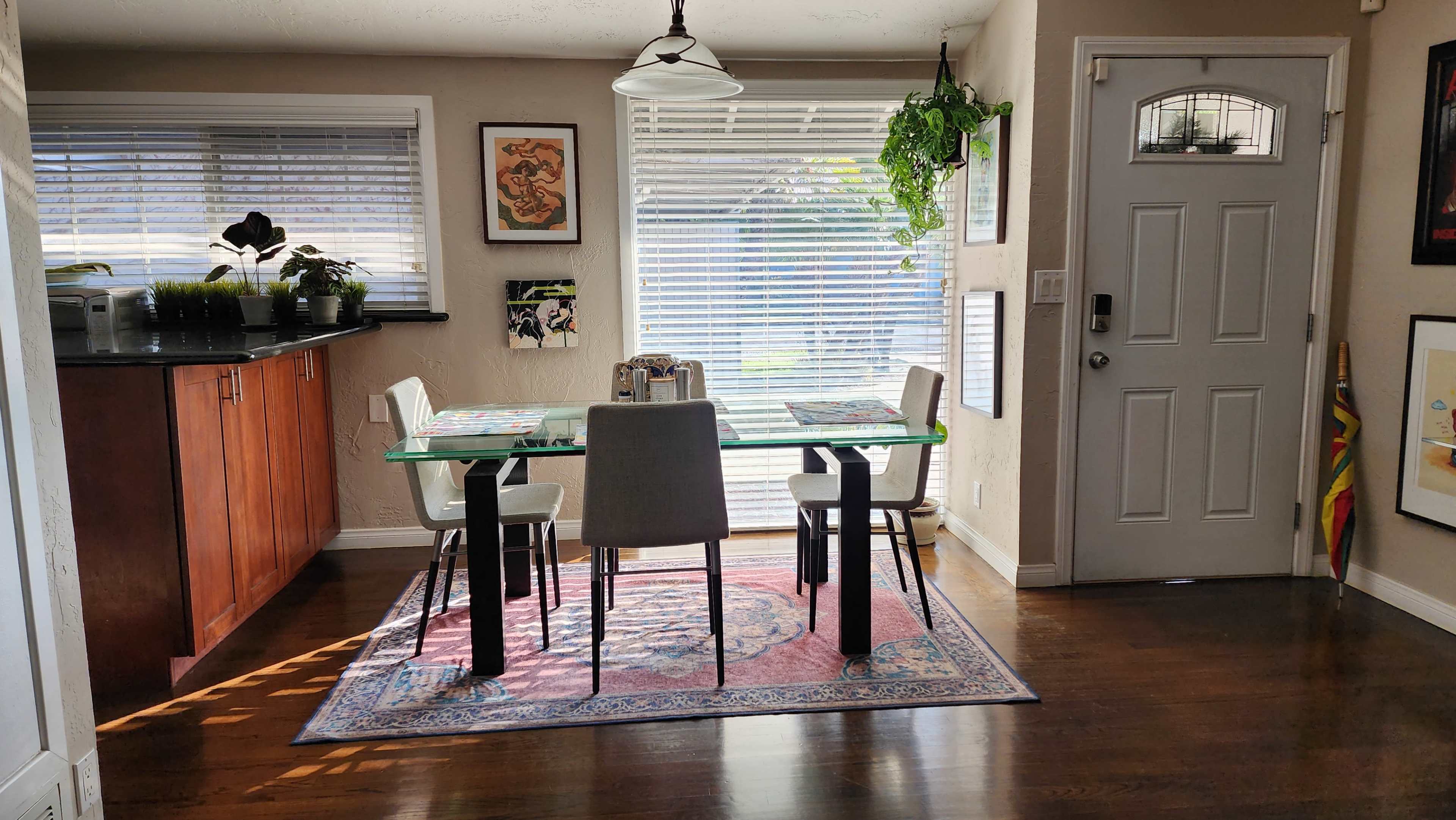 A dining area features a glass table with four chairs, surrounded by large windows allowing natural light, and a door leading outside.