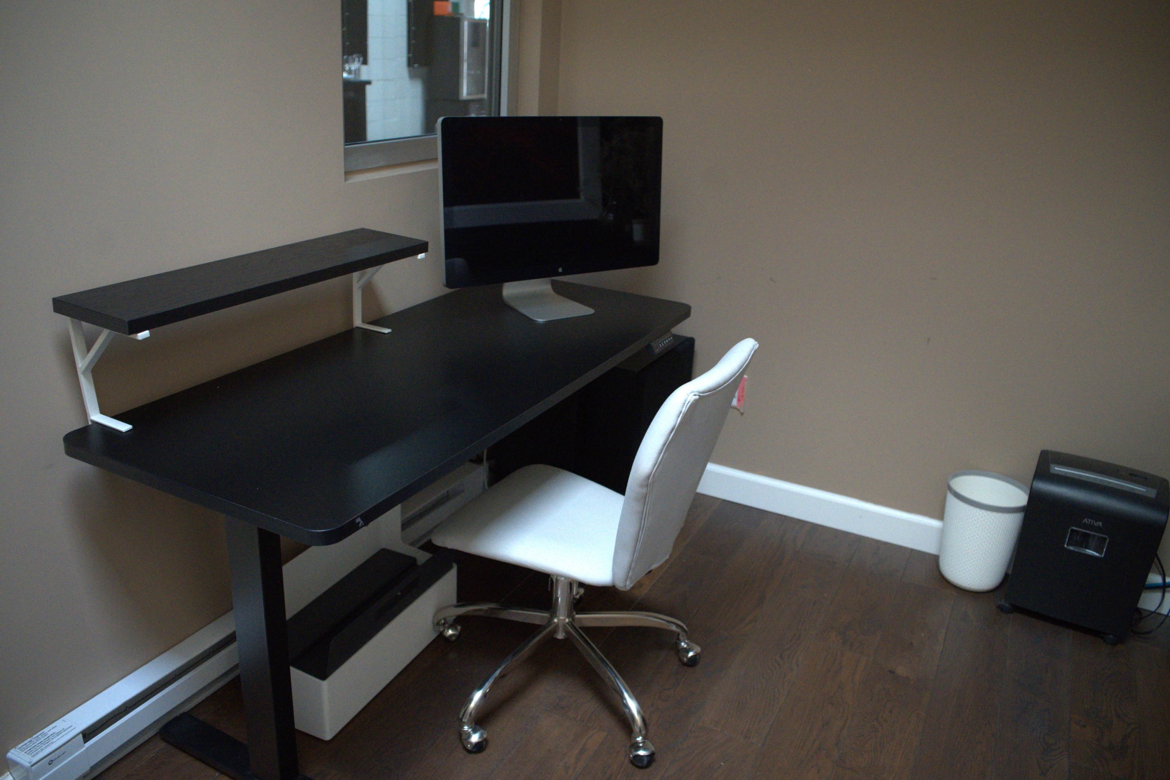 The image shows a minimalist workspace featuring a black desk with a monitor on a stand, a white office chair, and a small wastebasket in the corner.