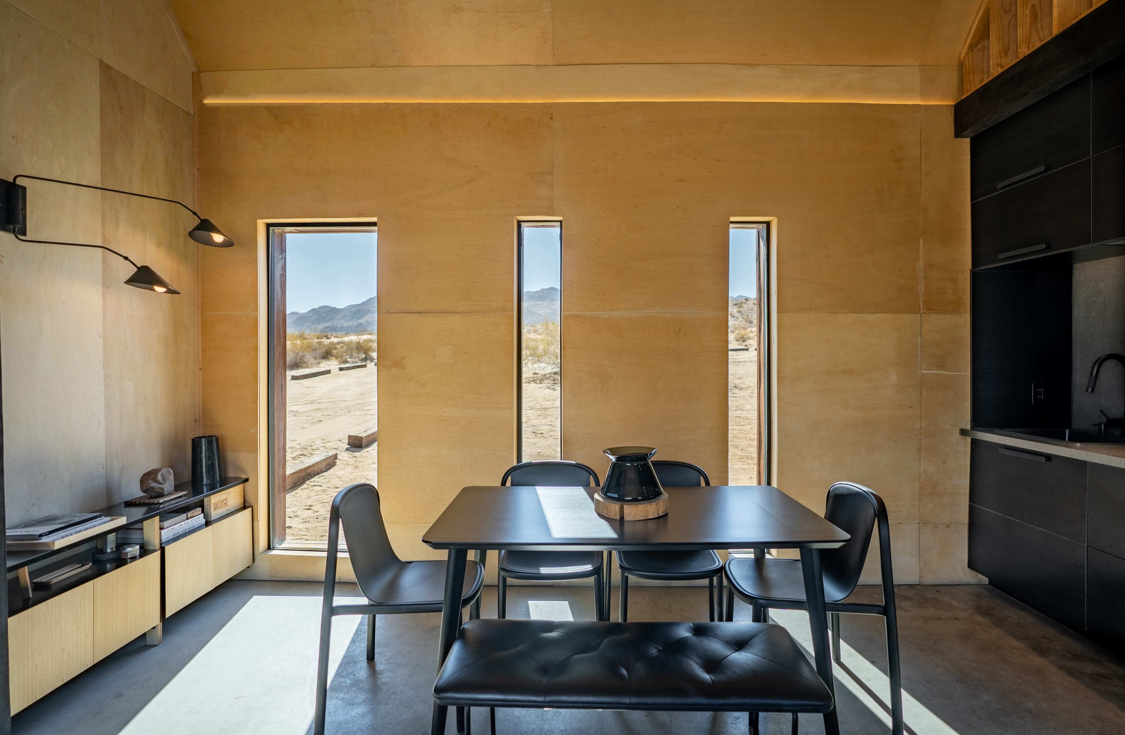 The interior of a minimalist room features a black table with four chairs, large windows allowing natural light, and a small kitchenette area against one wall.
