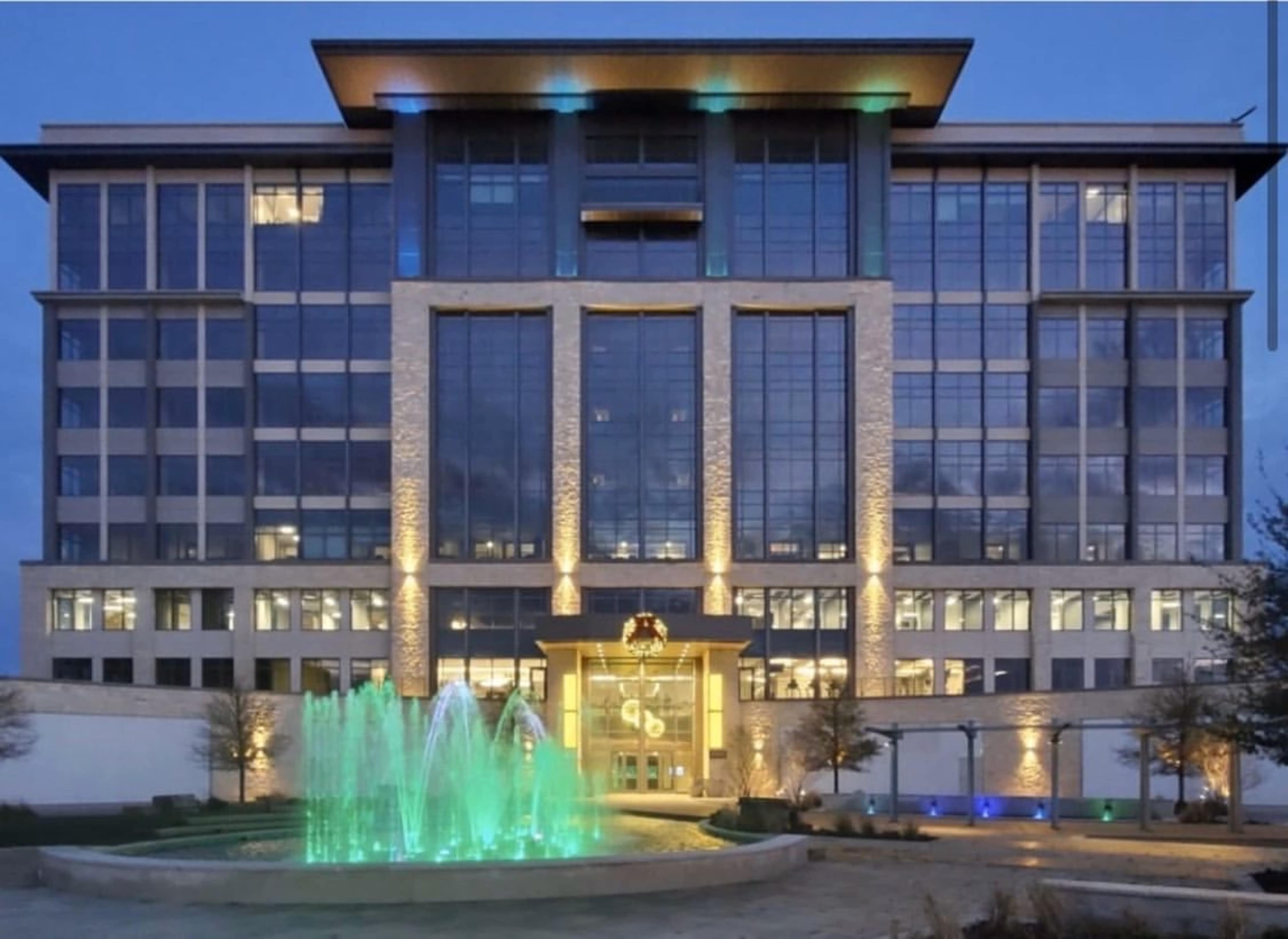 The image shows a modern multi-story building with large glass windows and a lit water fountain in front, illuminated at dusk.