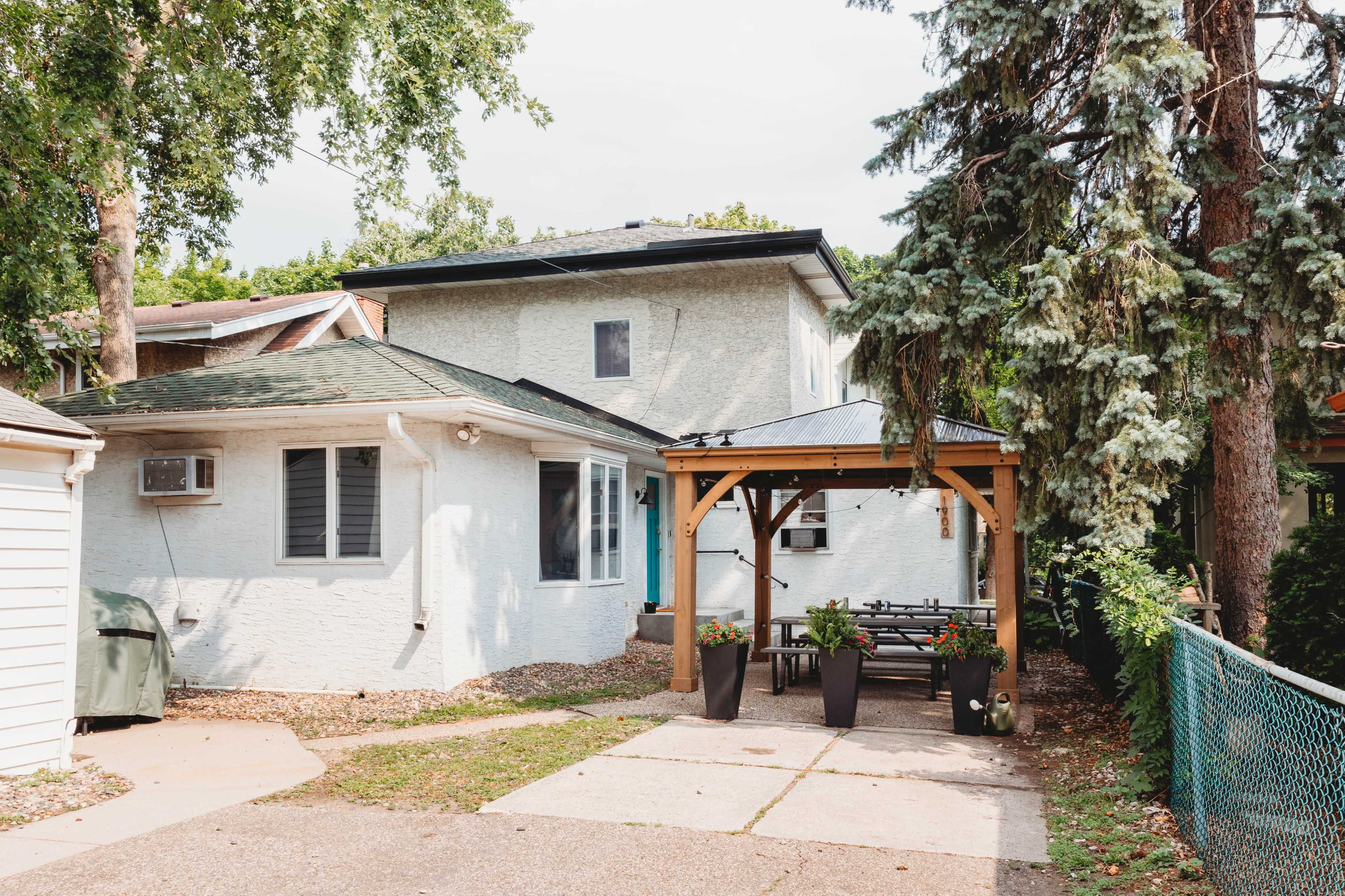 A two-story, light-colored house is shown with a paved driveway, a covered outdoor seating area, and trees alongside the property.