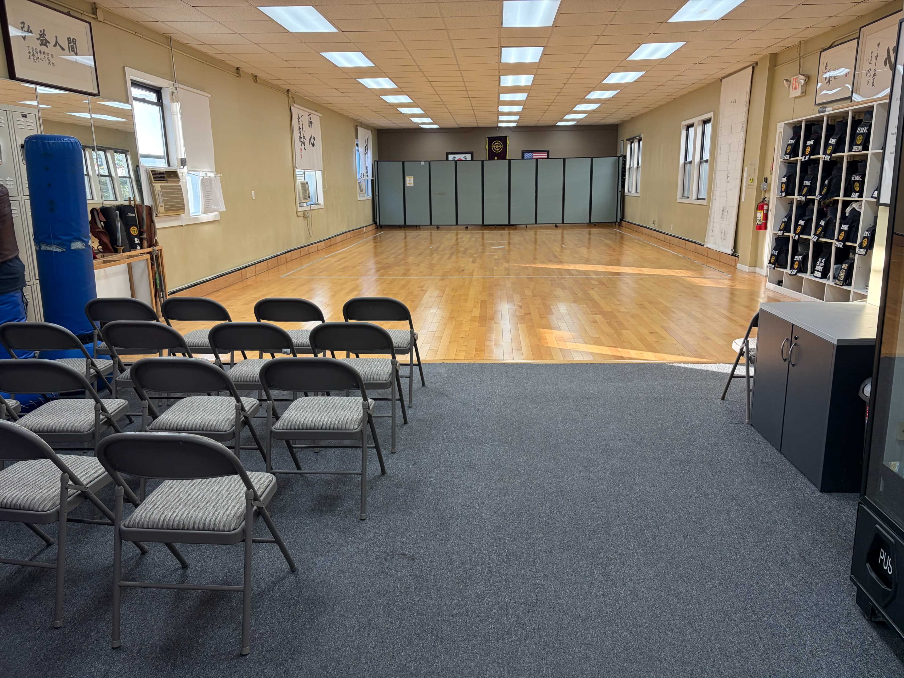 The image shows a spacious training room with wooden flooring, a row of empty chairs in the foreground, and divided sections in the background.
