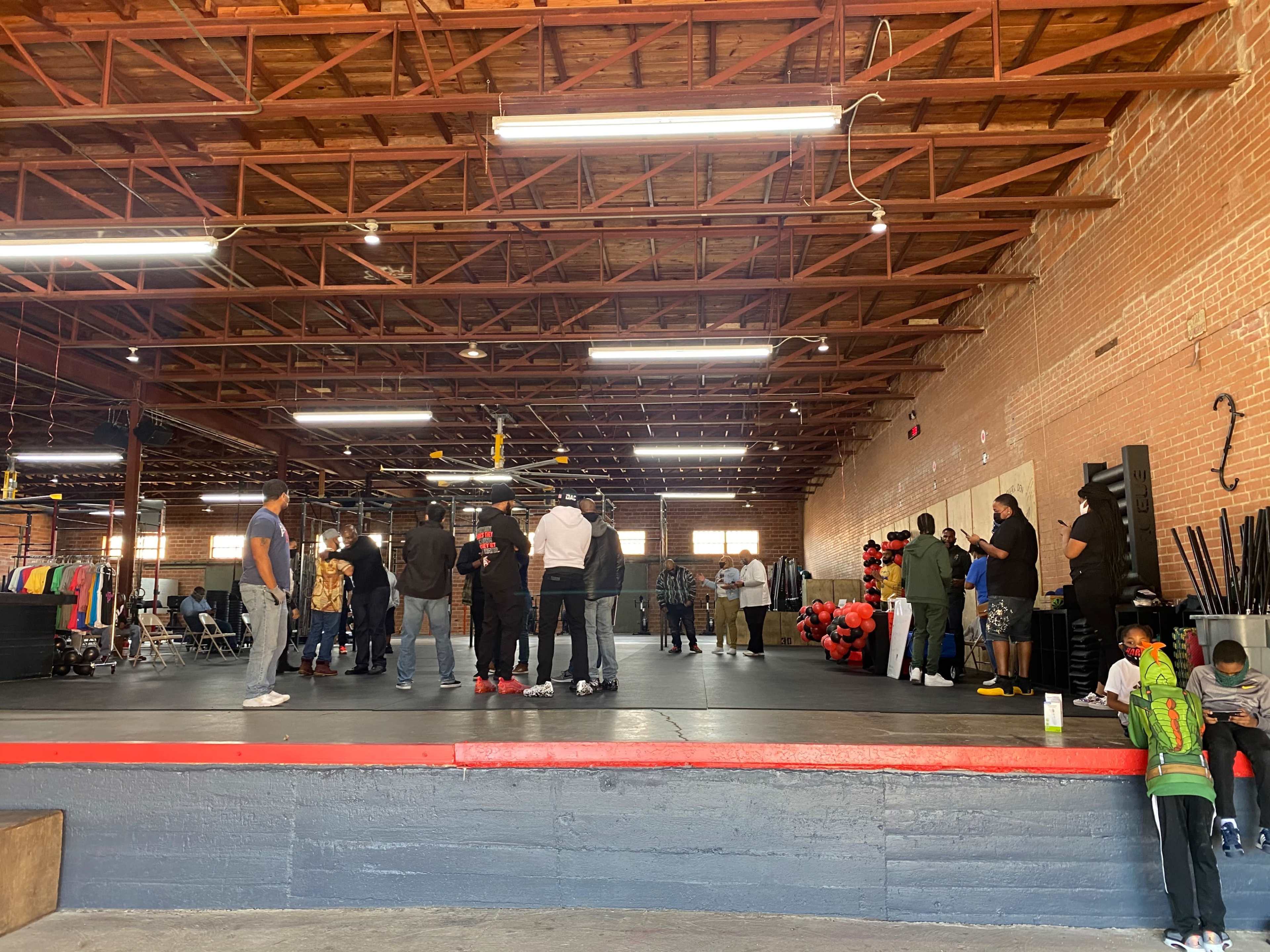 A group of people gathers in a spacious, well-lit warehouse with a wooden ceiling and red and black decorations.