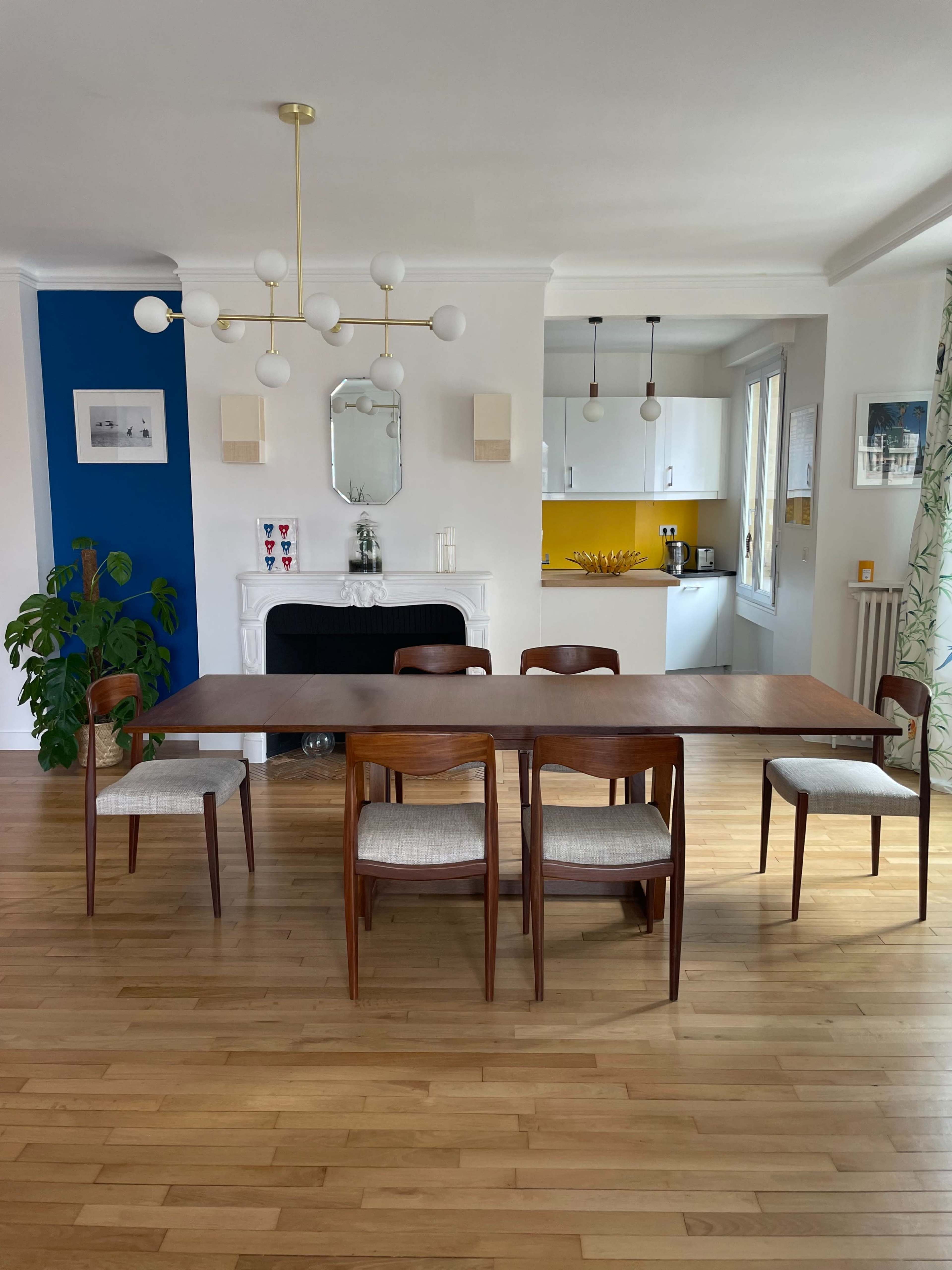 A wooden dining table is surrounded by four chairs in a bright room featuring a blue accent wall and a kitchen area visible in the background.