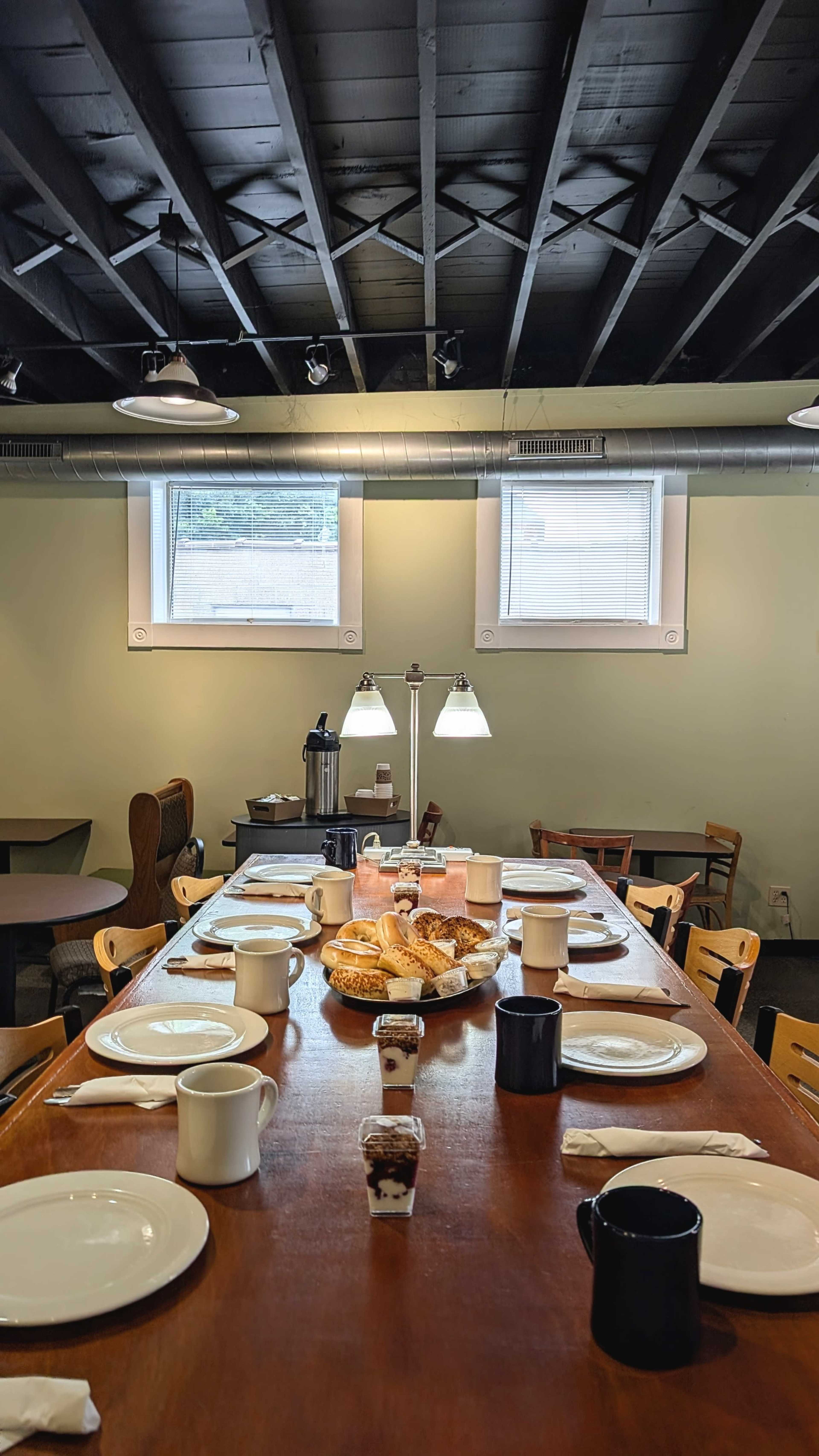 A long wooden dining table is set with plates, cups, and pastry items, illuminated by two lamps in a room with green walls and a black ceiling.