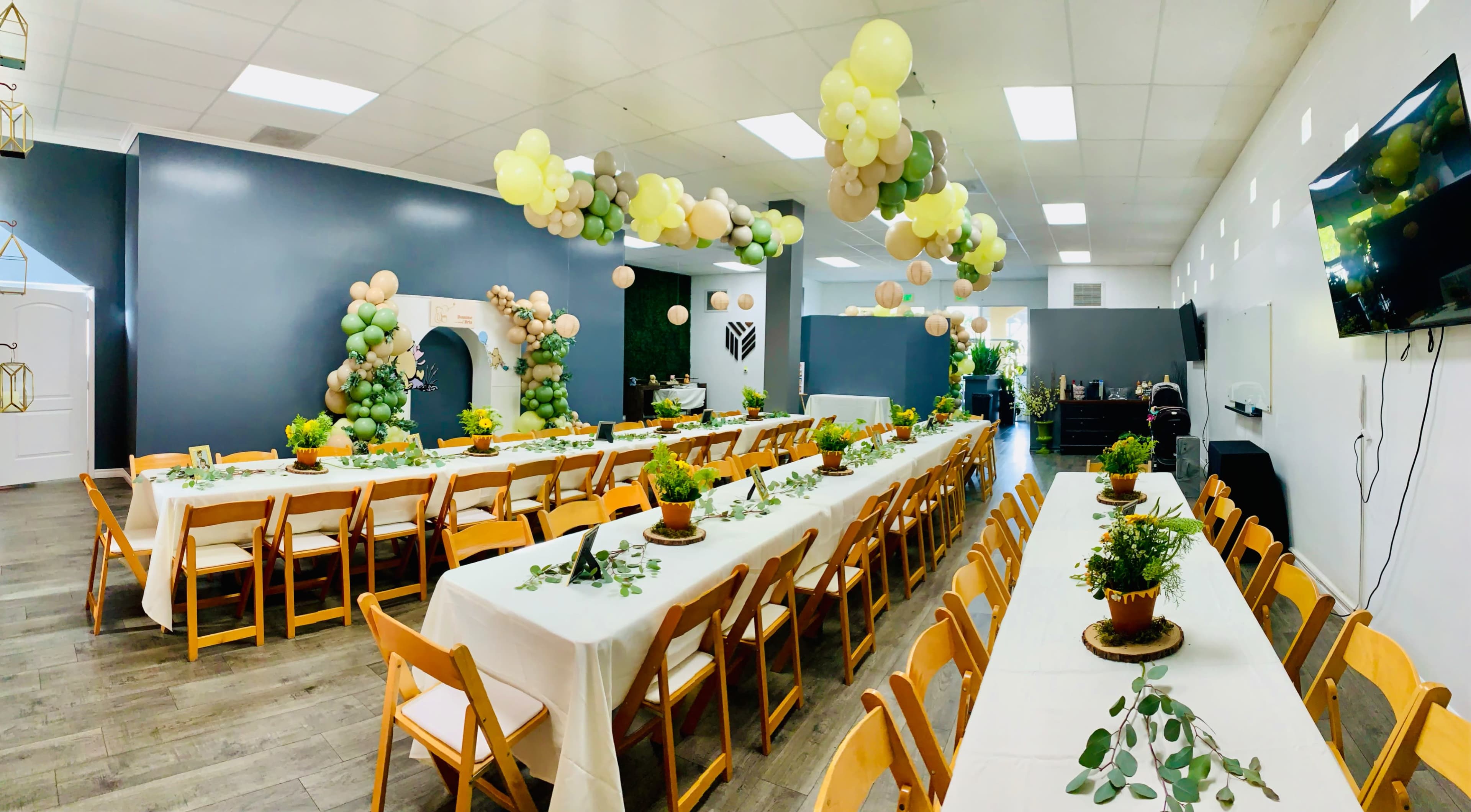 A spacious banquet hall is set up with long tables decorated with potted plants and balloons hanging overhead.