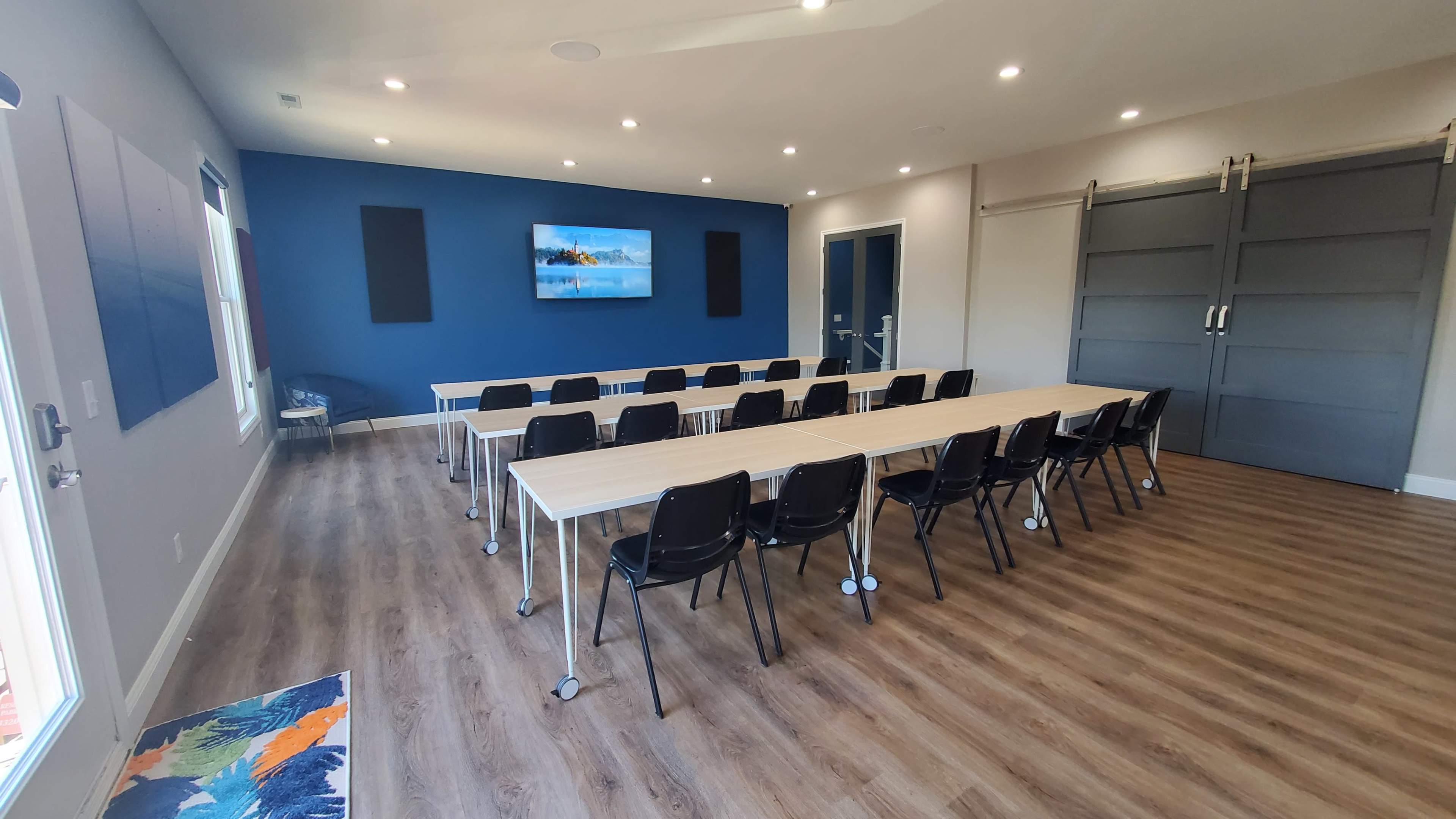 A modern meeting room features long tables arranged in rows facing a large screen on the wall, with a blue accent wall and large windows.