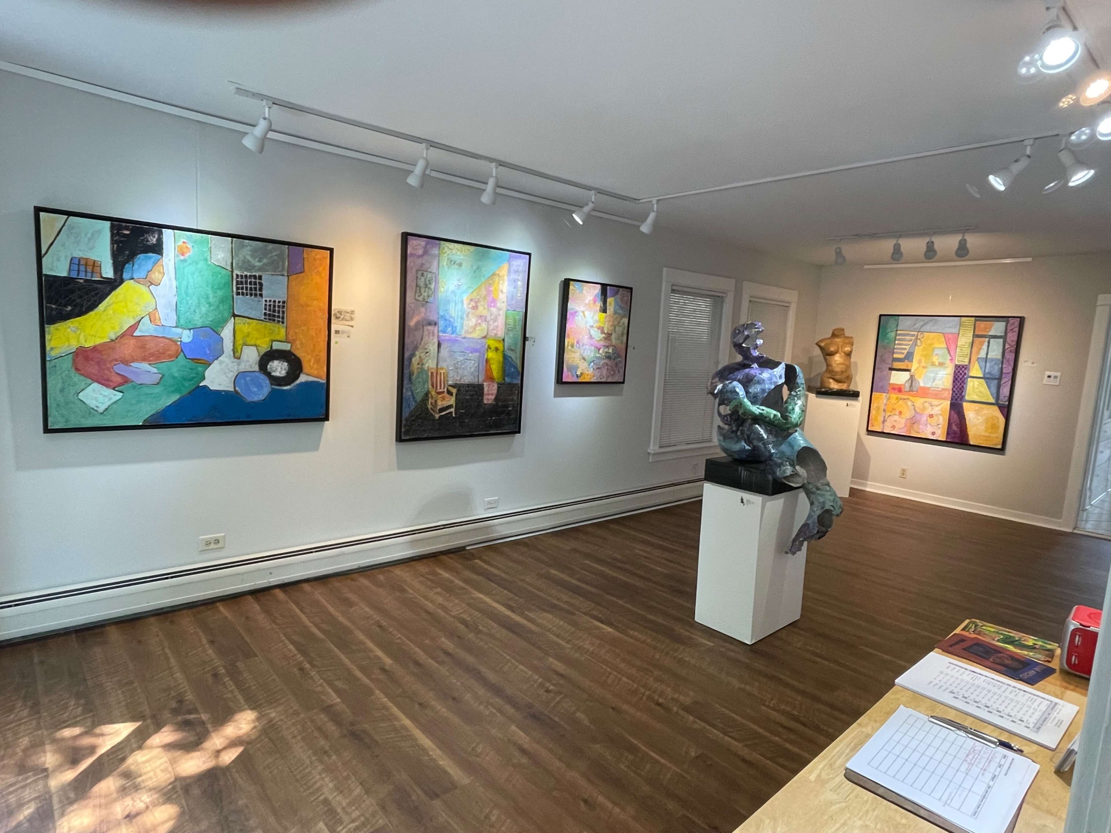An art gallery featuring various colorful paintings on the walls and two sculptures displayed on pedestals.