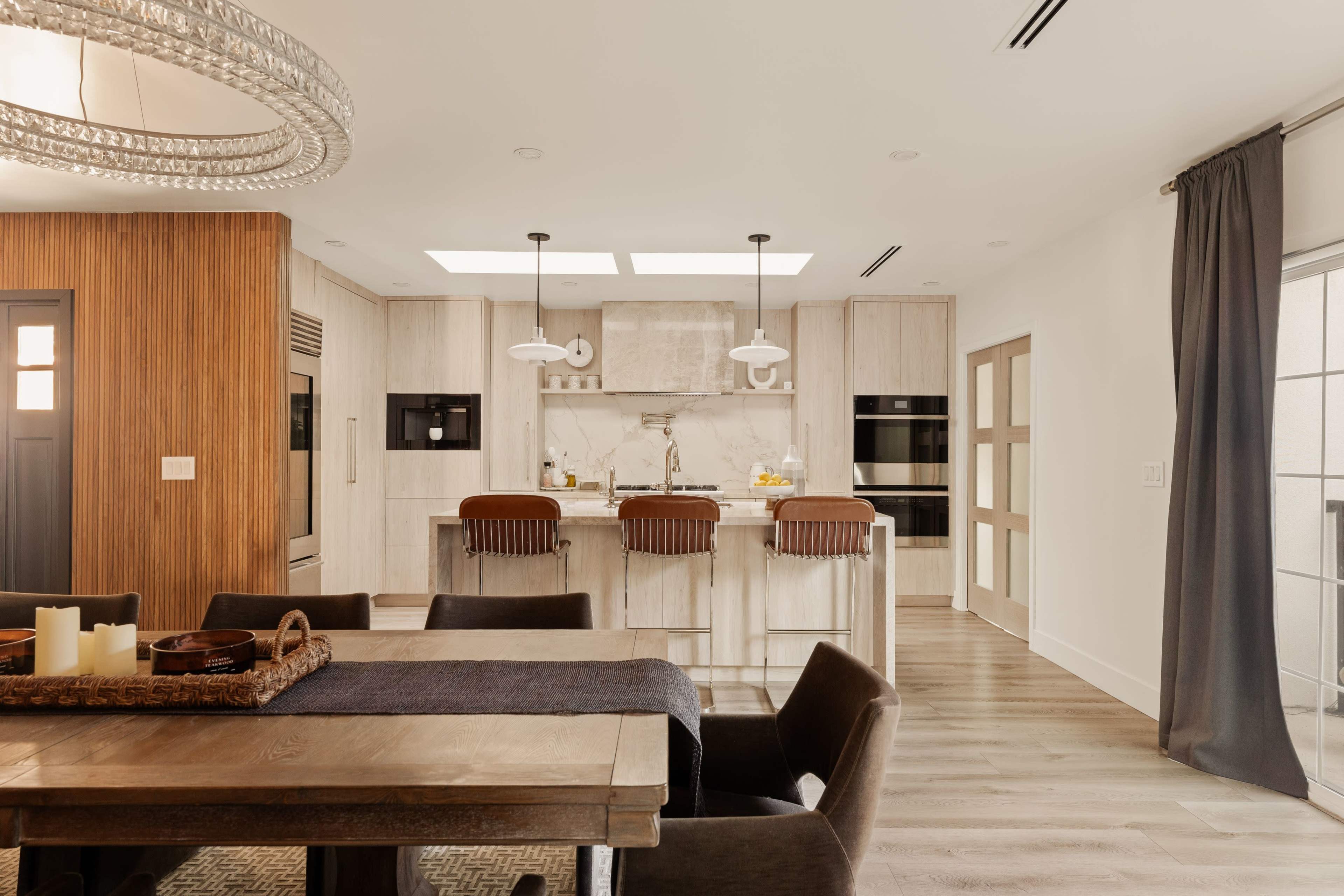 The image shows a modern kitchen and dining area with a wooden dining table, stylish seating, and a kitchen island featuring pendant lighting and a marble backsplash.