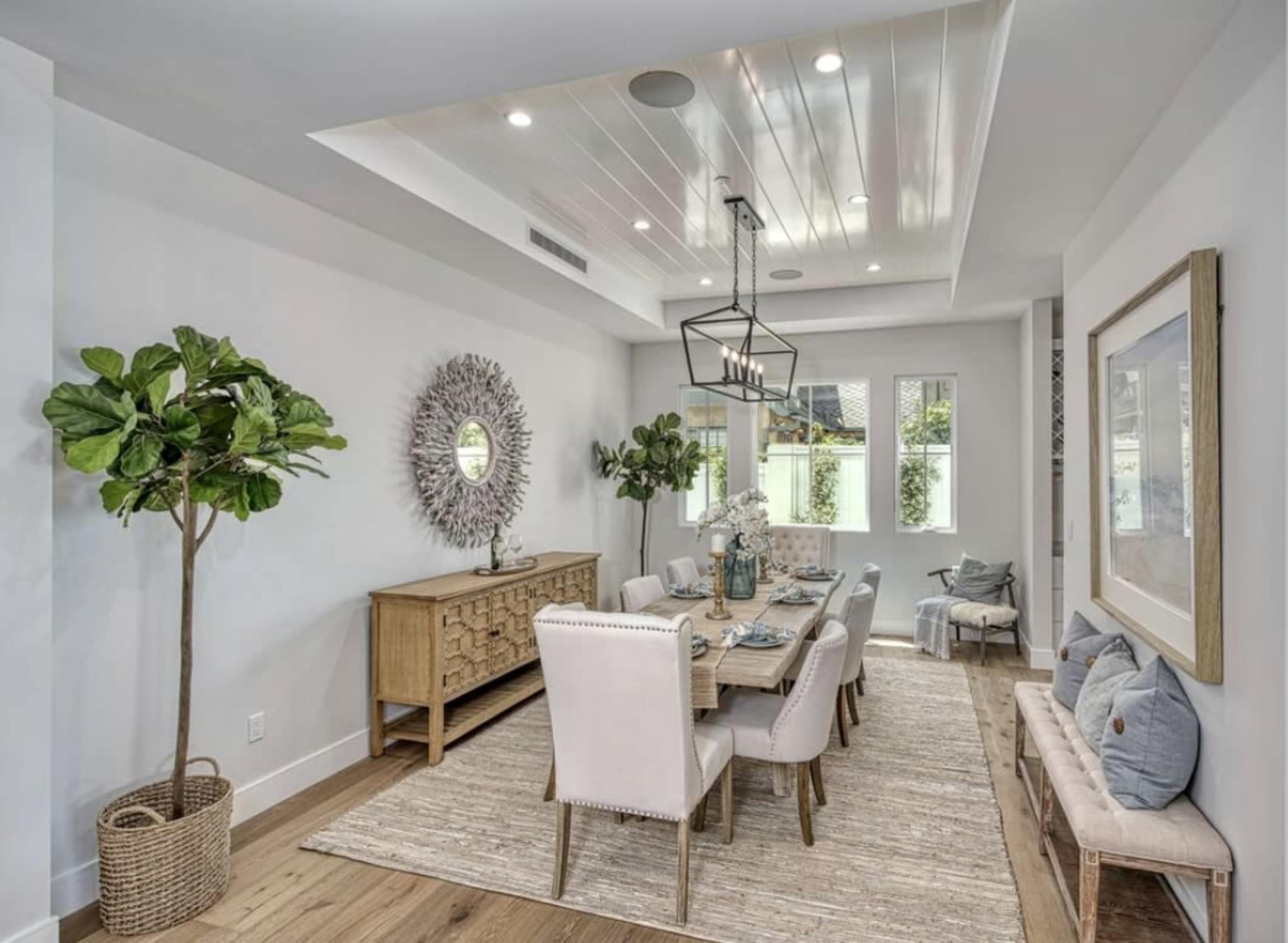 A modern dining room features a long table surrounded by upholstered chairs, with a decorative mirror on the wall and large windows letting in natural light.