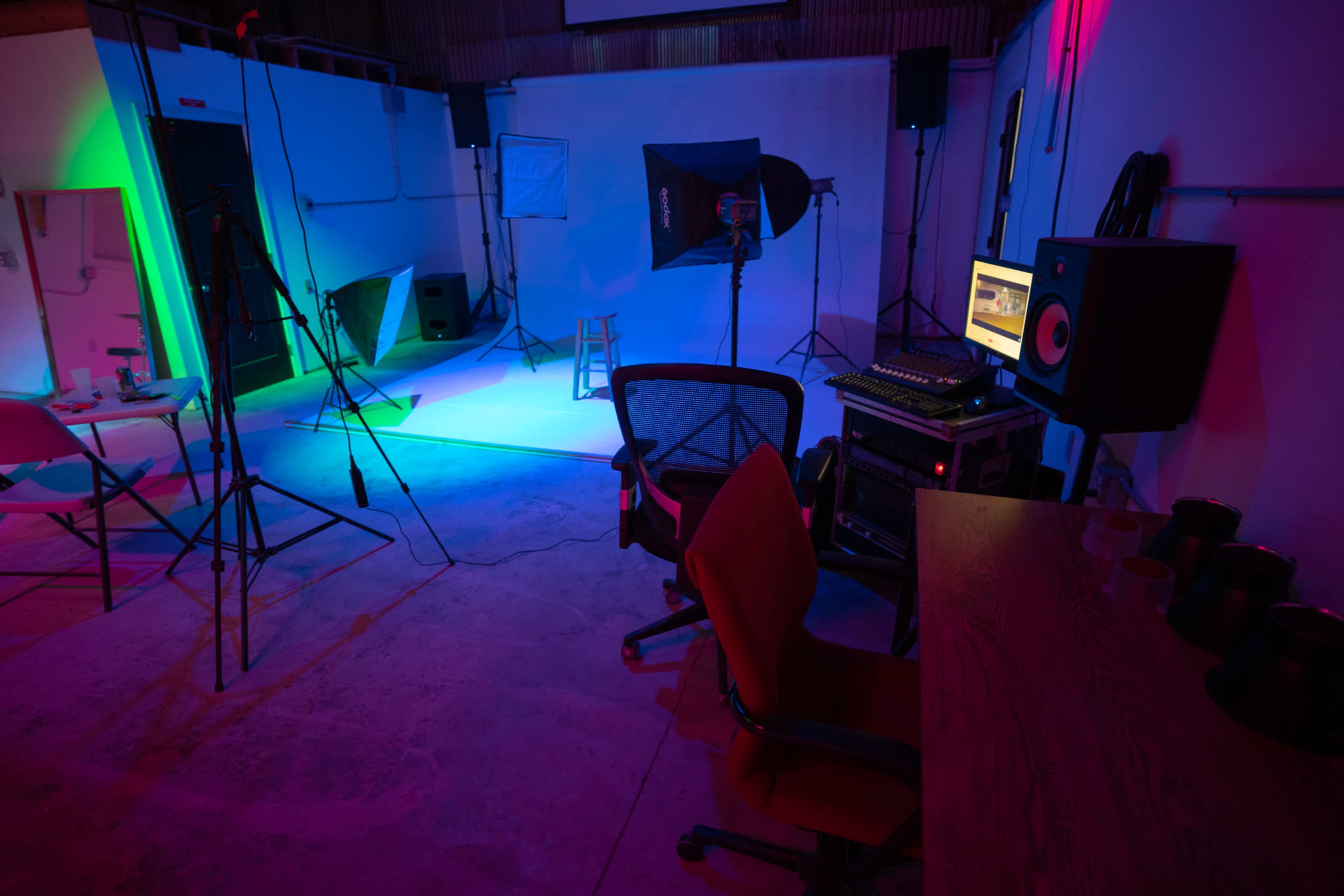 A photography studio setup with colorful lighting, a tall stool, a microphone stand, a computer monitor, and various equipment arranged around the space.