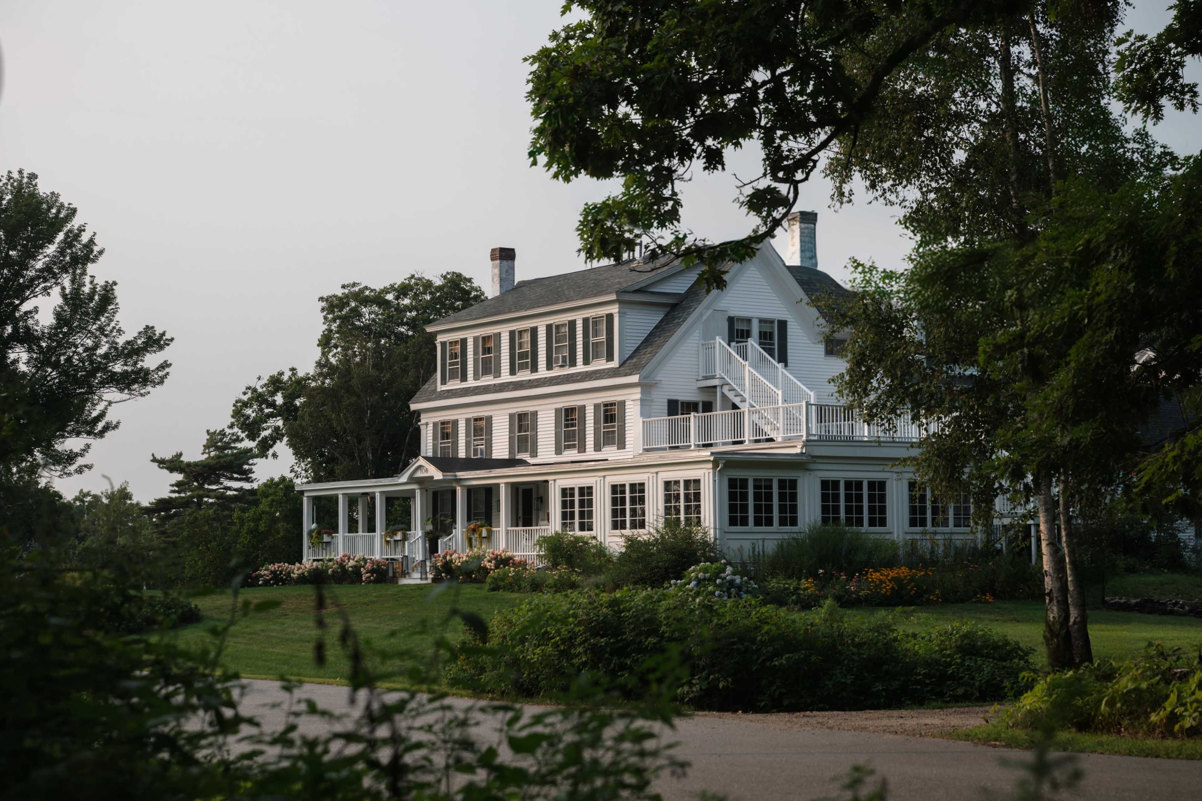 A large two-story white house with a porch and multiple windows is surrounded by green lawns and garden beds.