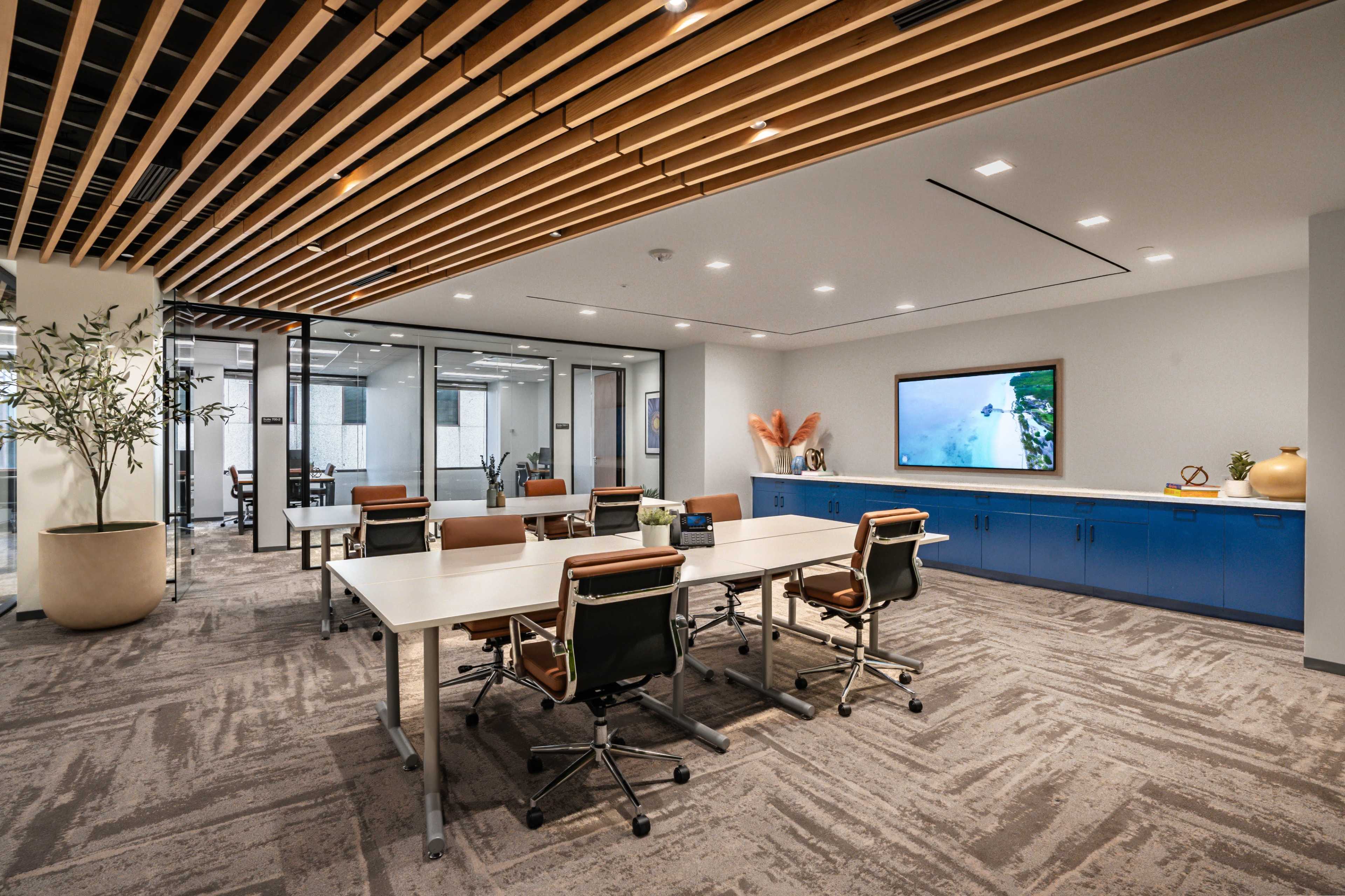 A modern office space features a series of desks arranged for collaboration, with glass partitions and a blue storage unit against a wall displaying a large screen.