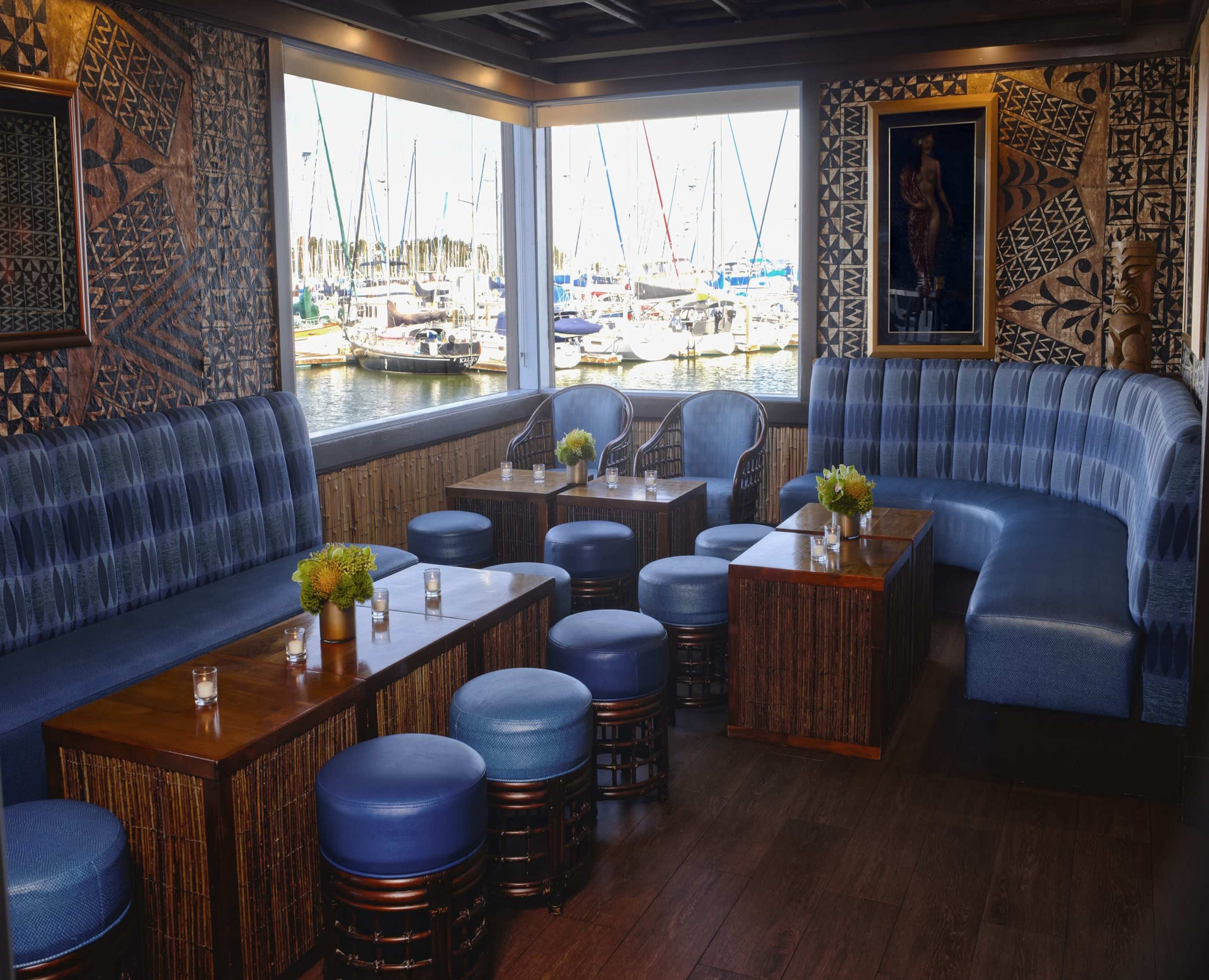 The image depicts a cozy waterfront lounge area featuring blue upholstered seating, wooden tables, and a view of sailboats through large windows.