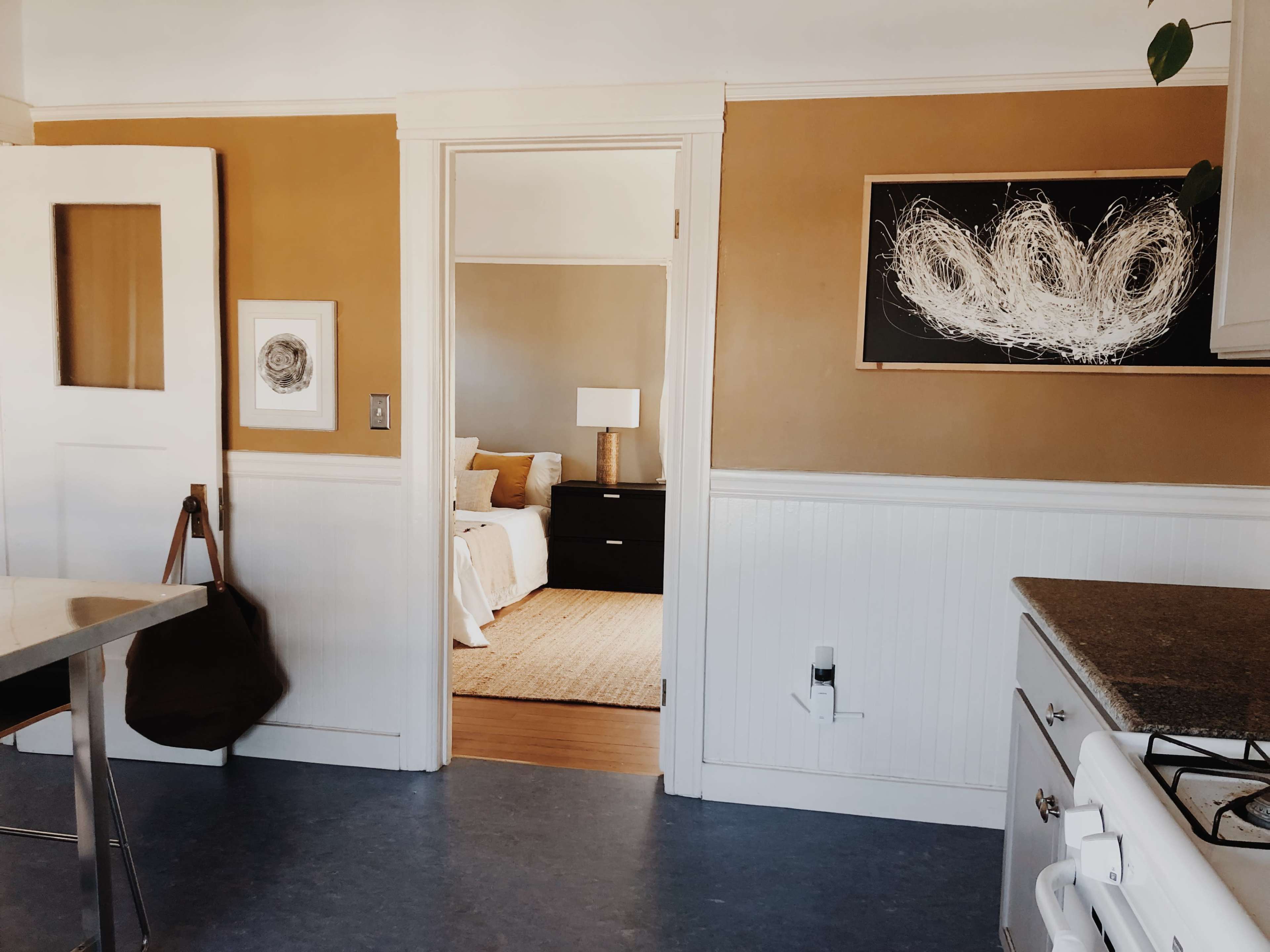 The image shows a kitchen with beige walls, a doorway leading to a bedroom, and an abstract artwork on the wall.