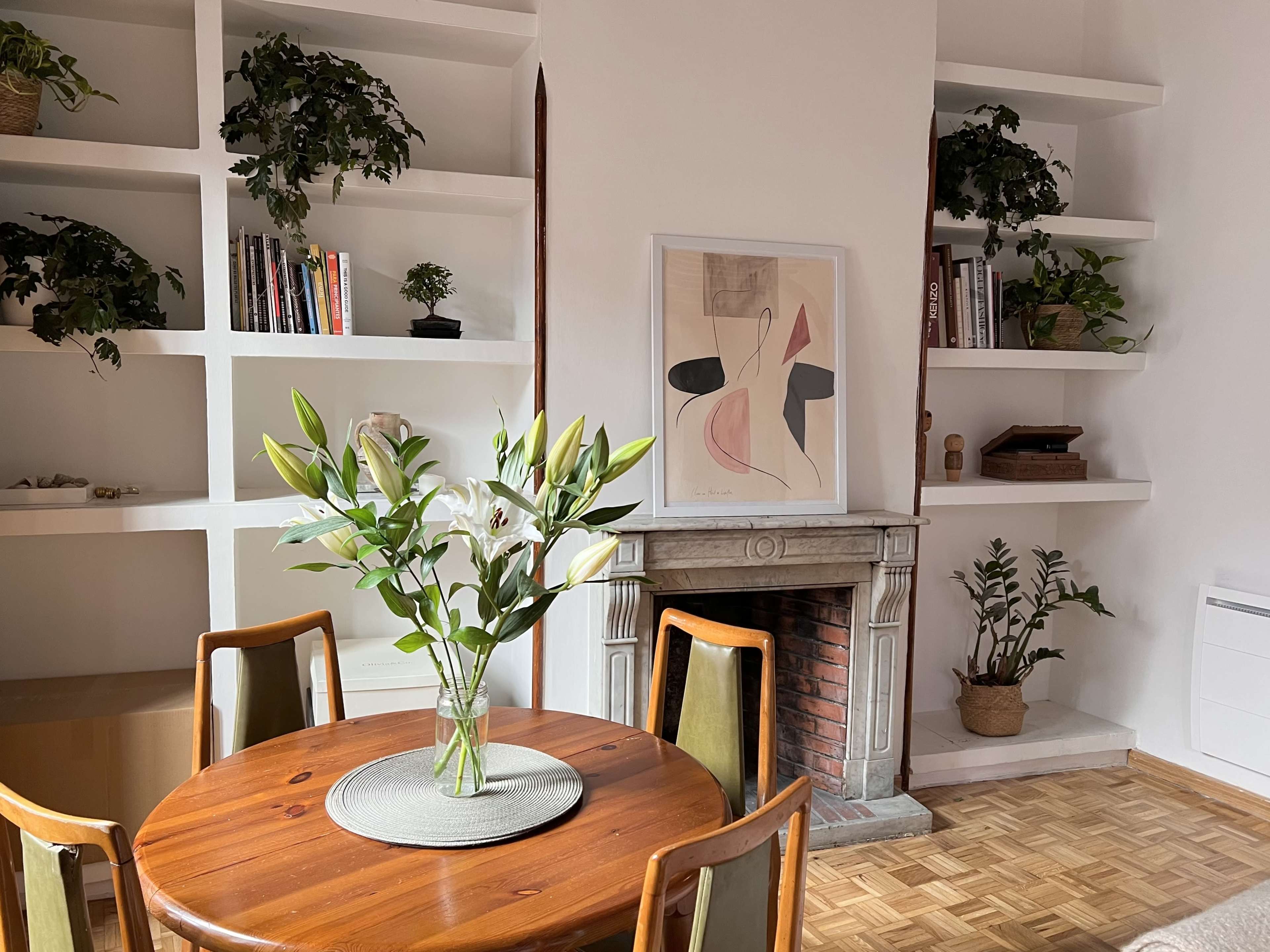 A round wooden dining table with a vase of lilies is positioned near a white wall with built-in shelves displaying plants and books.