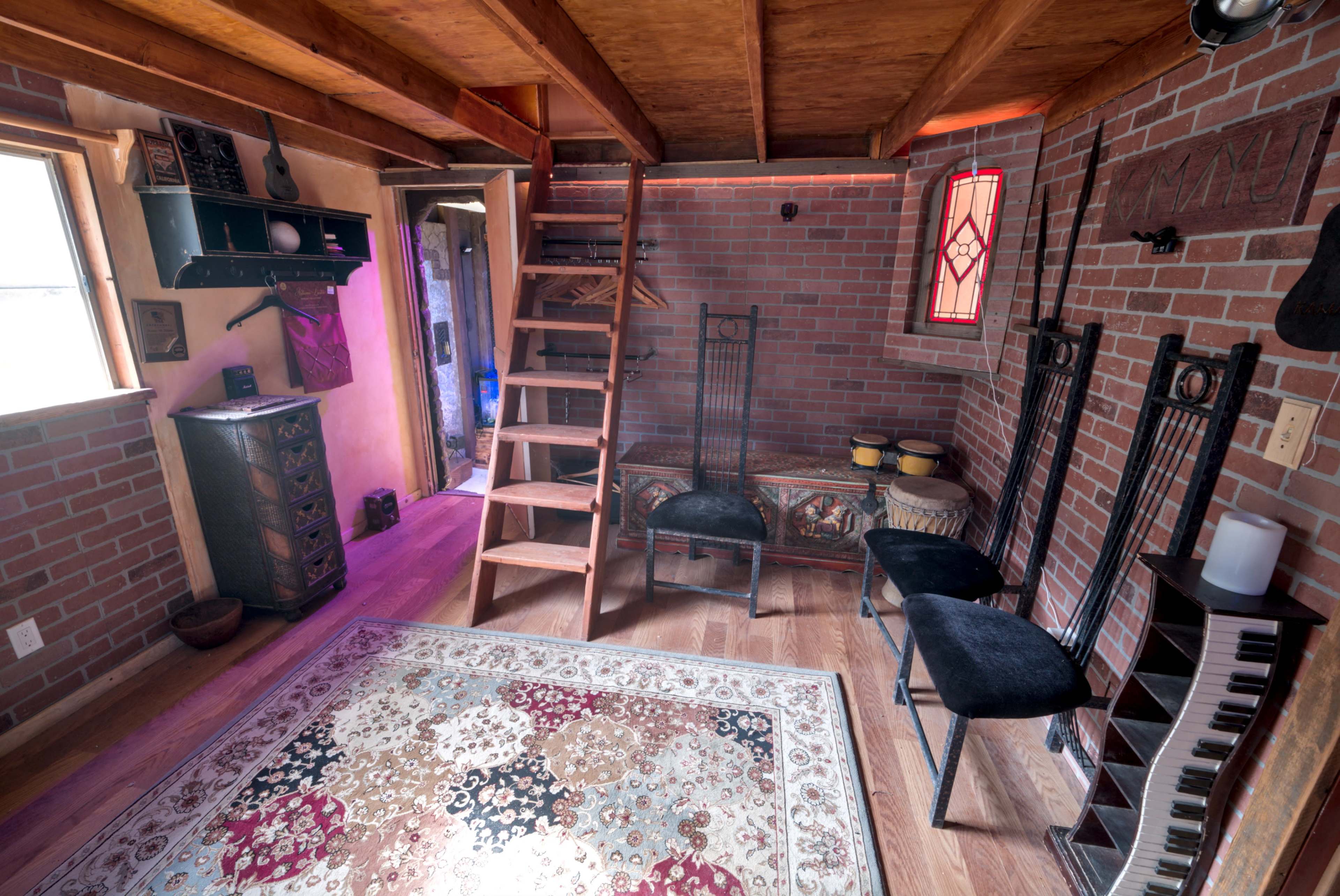 The interior of a cozy room features exposed brick walls, wooden beams, a staircase, and several chairs along with a colorful area rug.